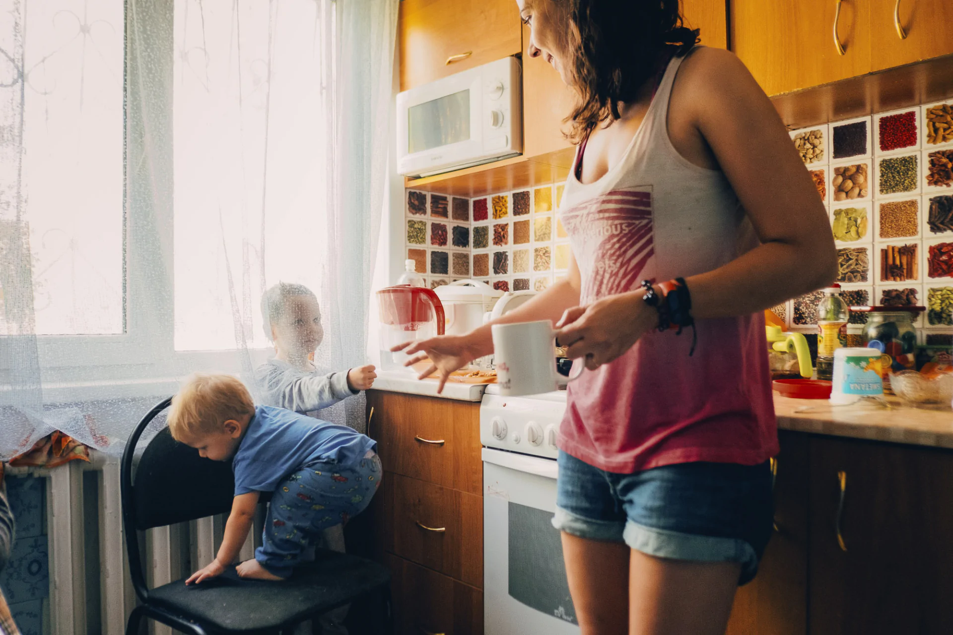 A woman in a kitchen with a child standing on a chair.