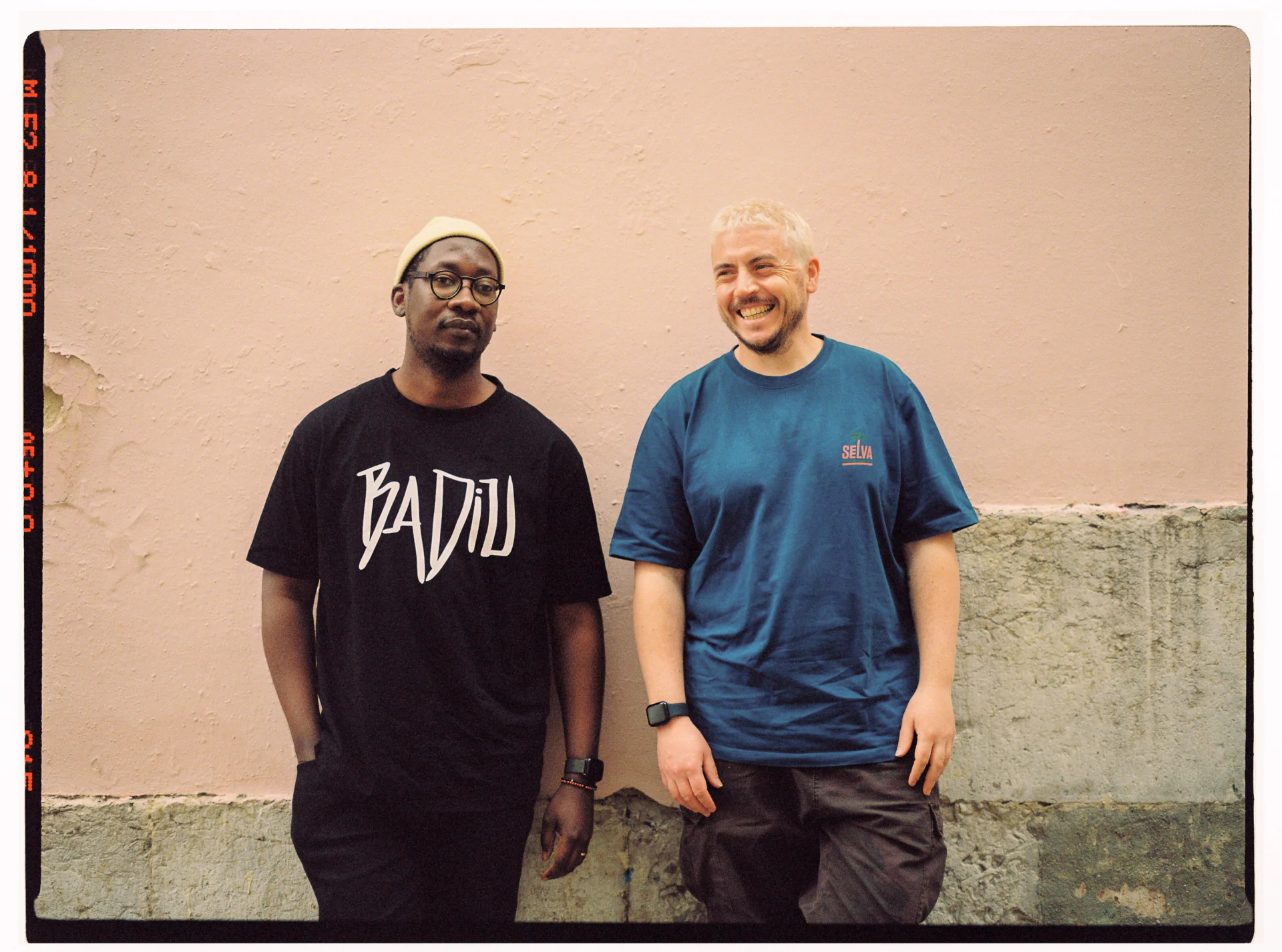 Two men standing next to each other in front of a pink wall.