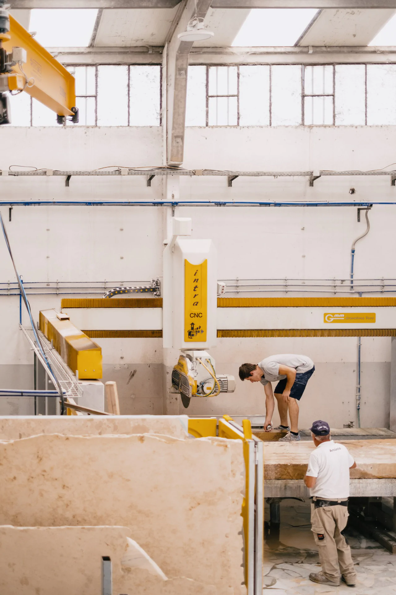 Two workers in a factory with one inspecting a large machine, while the other stands nearby. Large stone slabs are present in the foreground.