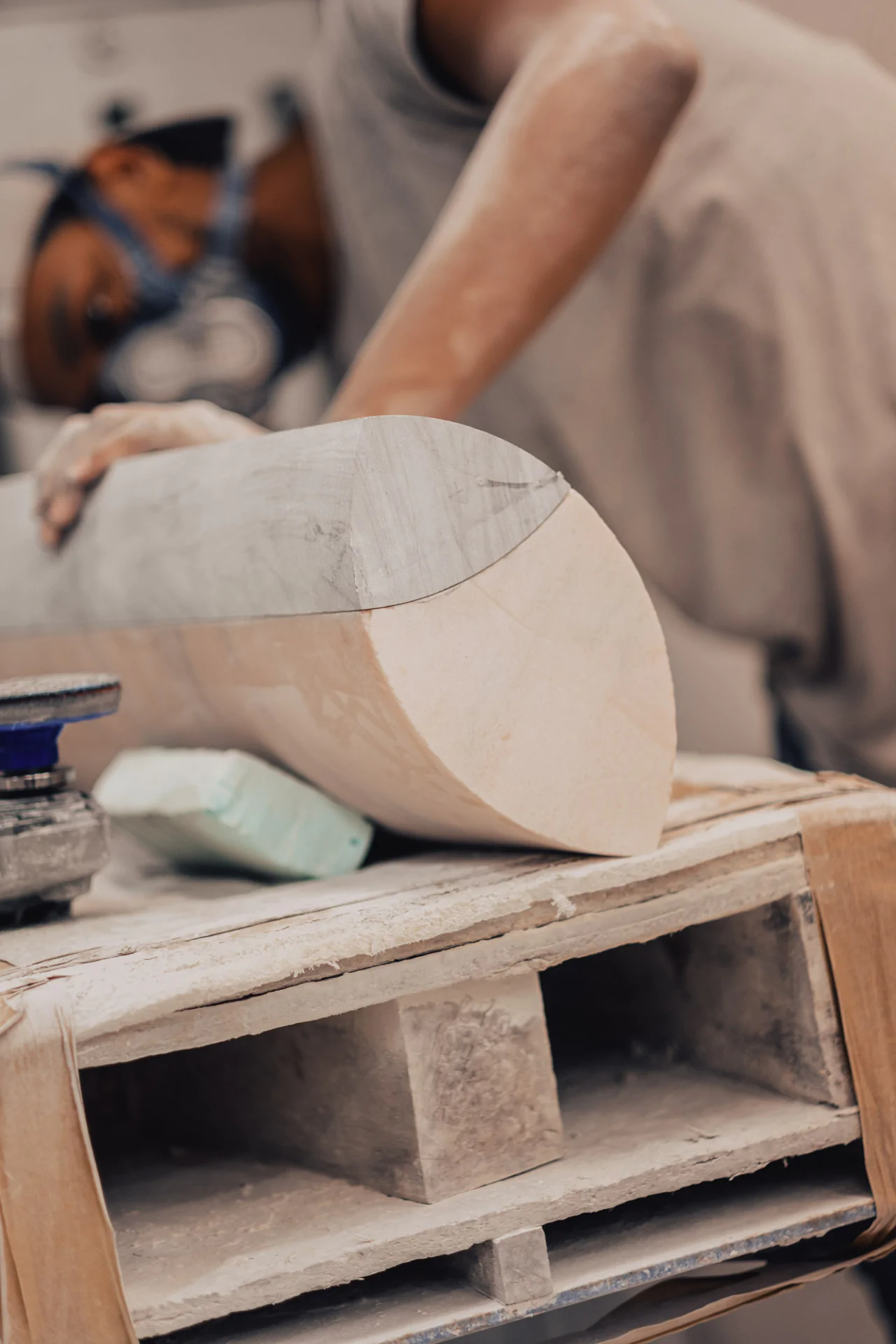 Person sanding a large piece of wood in a workshop, wearing a protective mask.