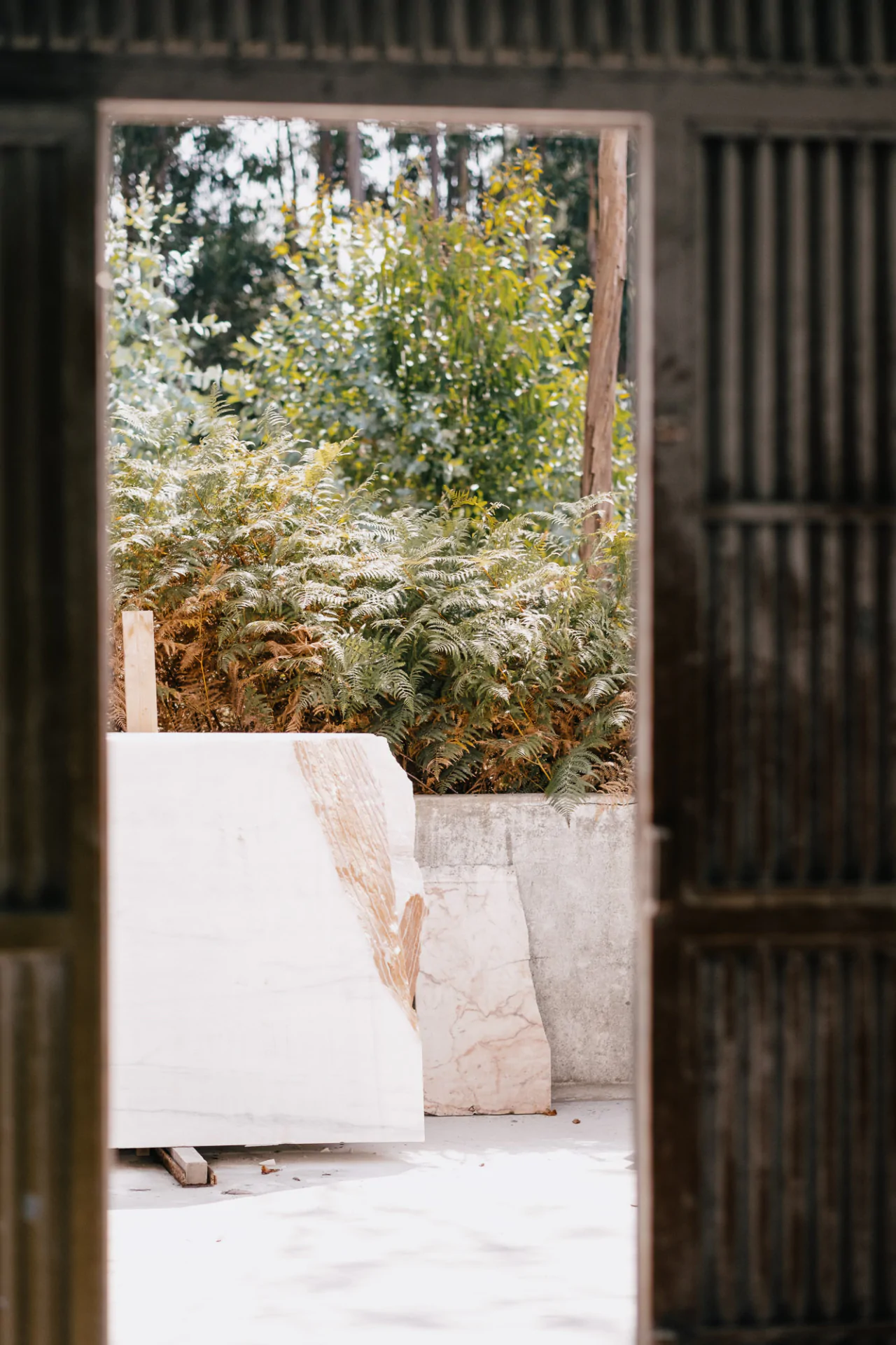 View through an open door of a patio with stone slabs leaning against a wall, surrounded by greenery and trees in the background.