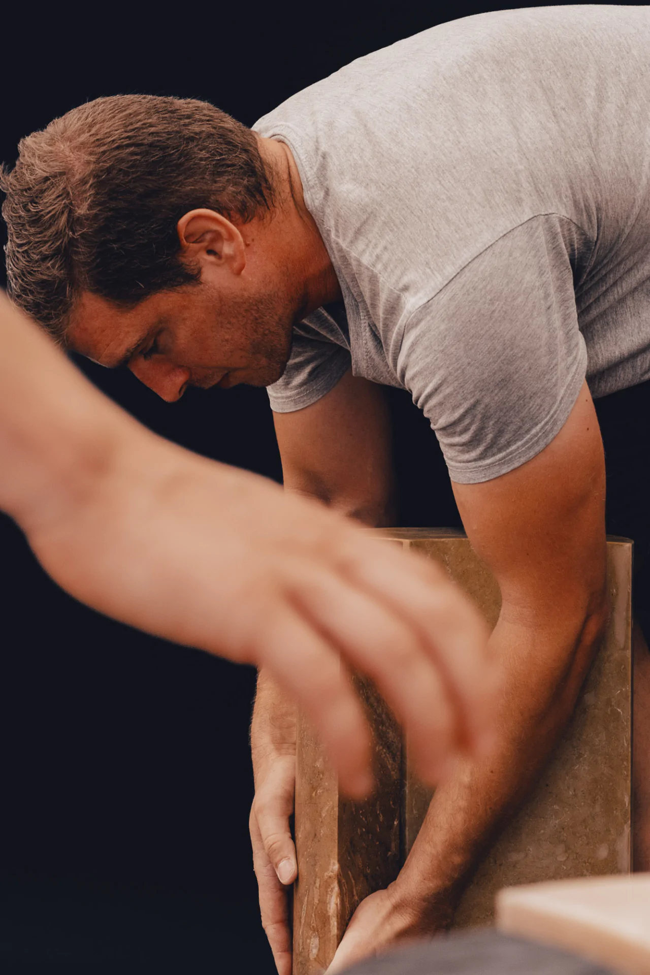 Person in a gray shirt bending down, adjusting or examining an object. Another hand in the foreground. Dark background.