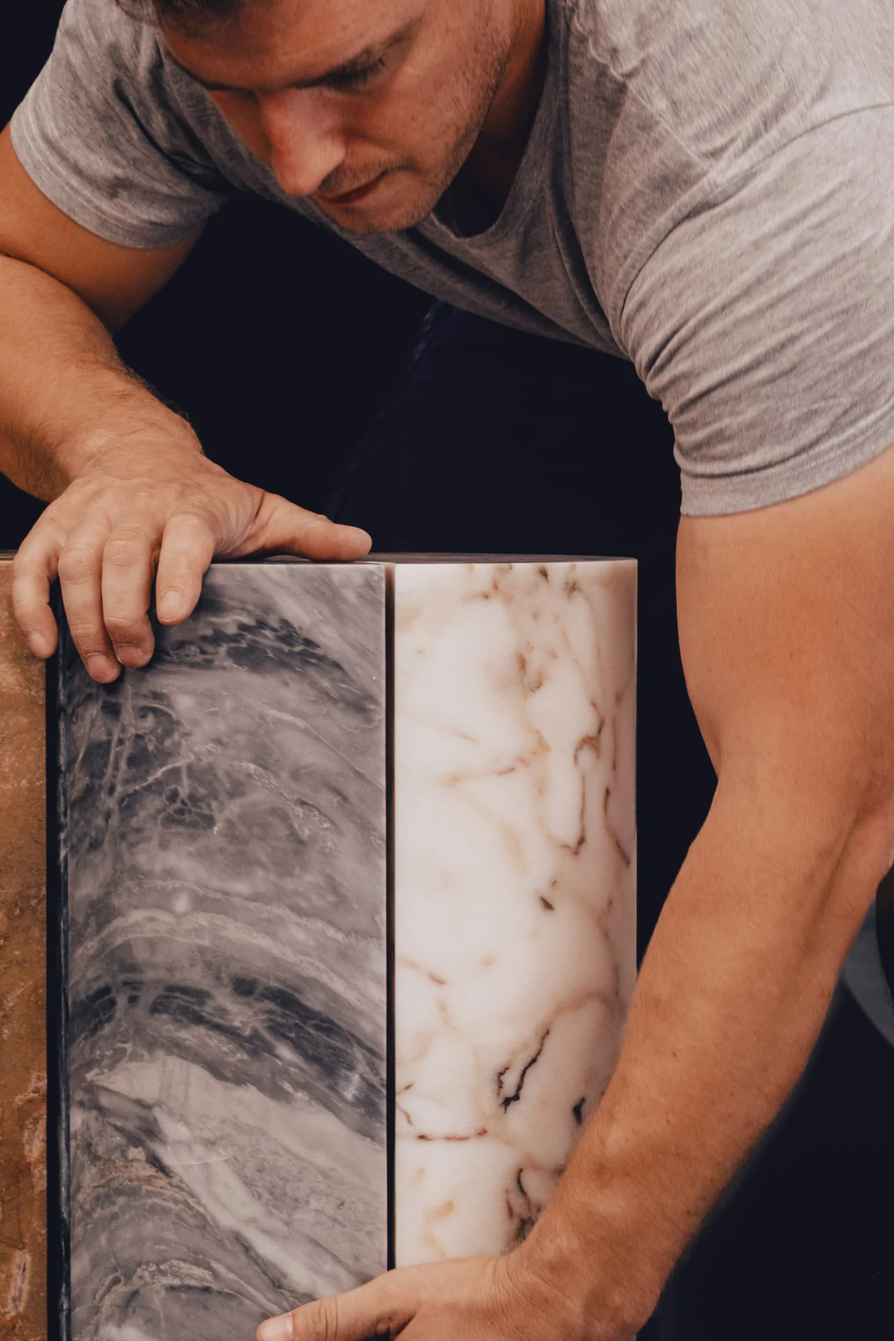 Person in a gray t-shirt examining a rectangular block made of gray and white marble sections, set against a dark background.