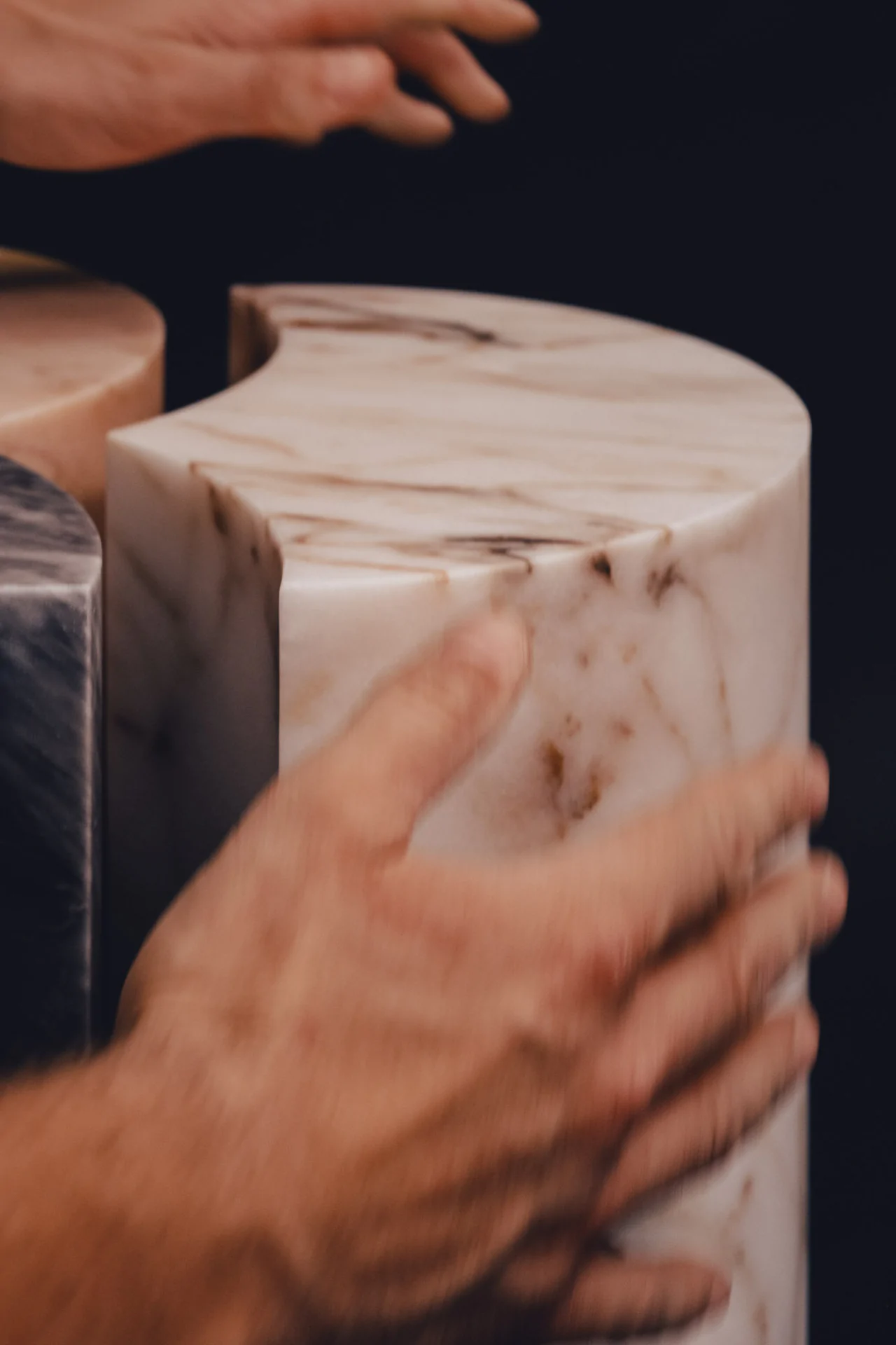 Hands touching smooth, cylindrical marble structures with varied colors on a dark background.