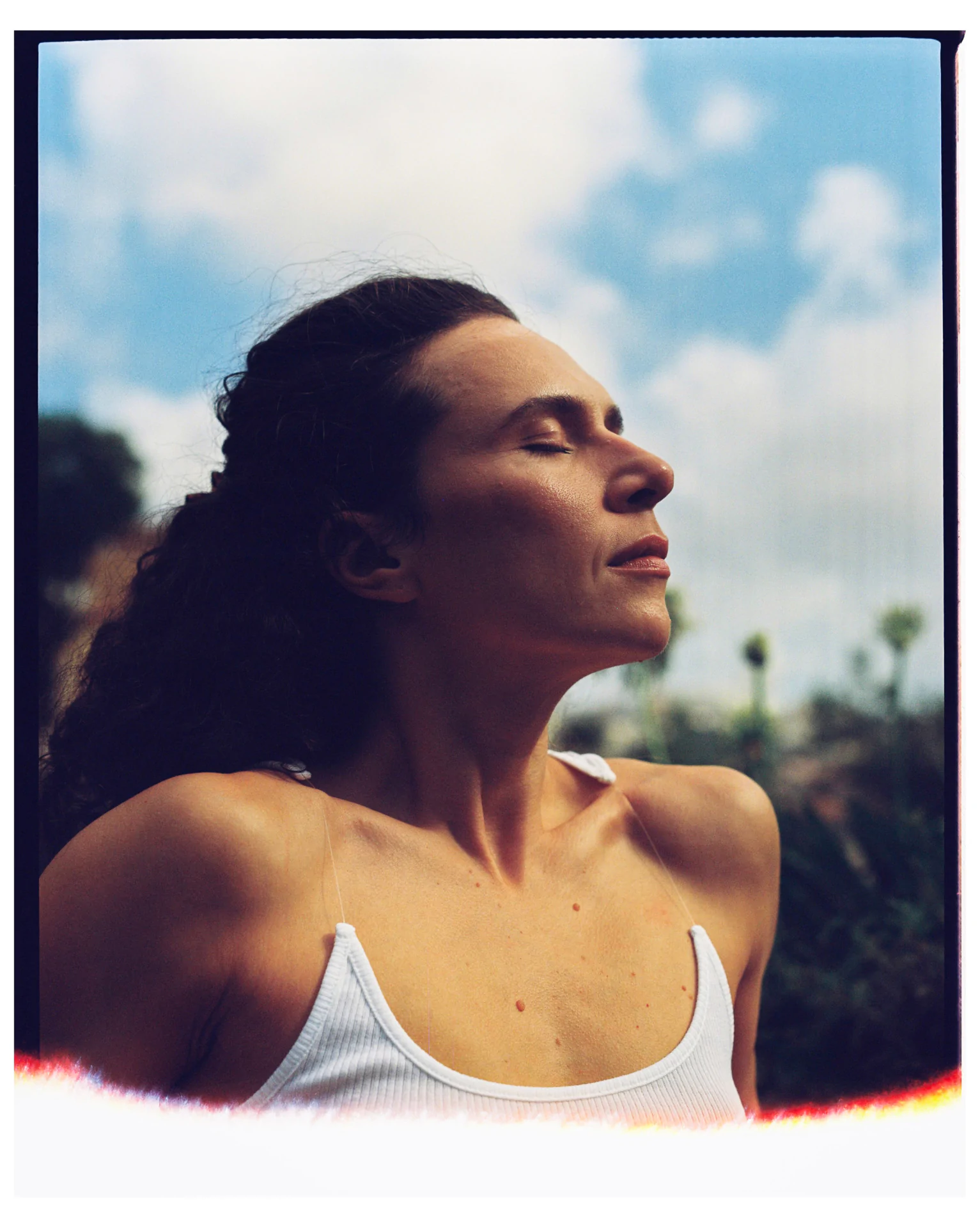 Woman in a white tank top stands outdoors with eyes closed, facing upwards under a partly cloudy sky.