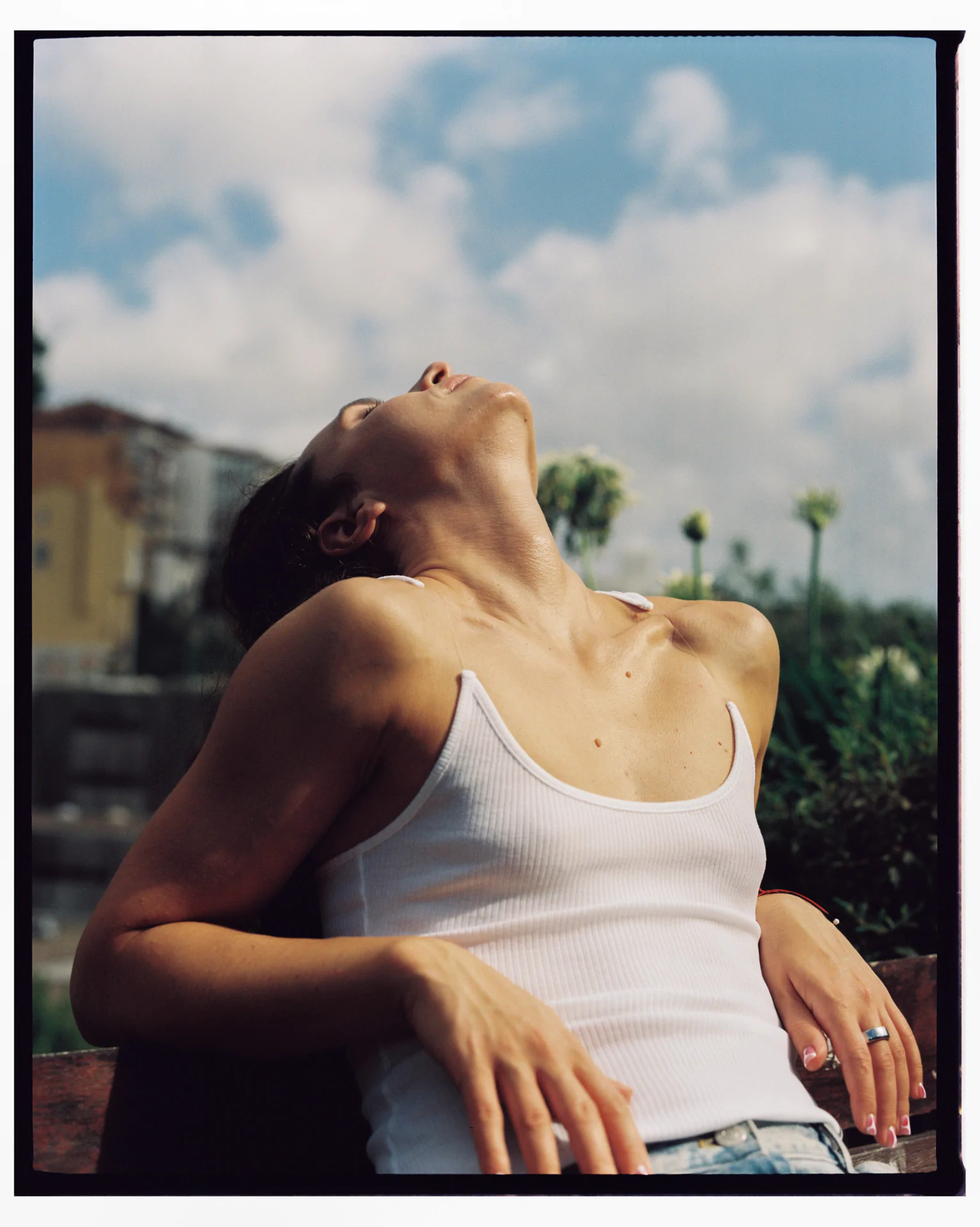 Person in a white tank top leans back on a bench, head tilted upward to the sky, with eyes closed. A blurred outdoor background features greenery and a partly cloudy sky.