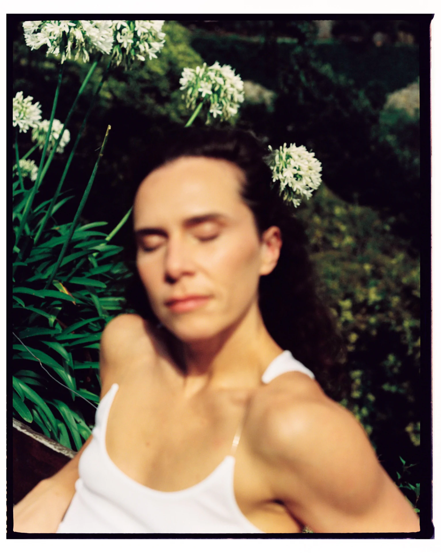 A person in a white tank top, out of focus, with closed eyes sits near green plants with white flowers in the background.