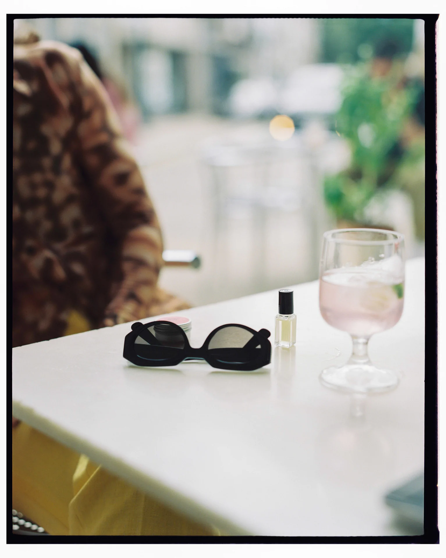 A pair of black sunglasses, a small bottle of perfume, and a glass filled with pink liquid on a white table. A person in patterned clothing is seated in the background.