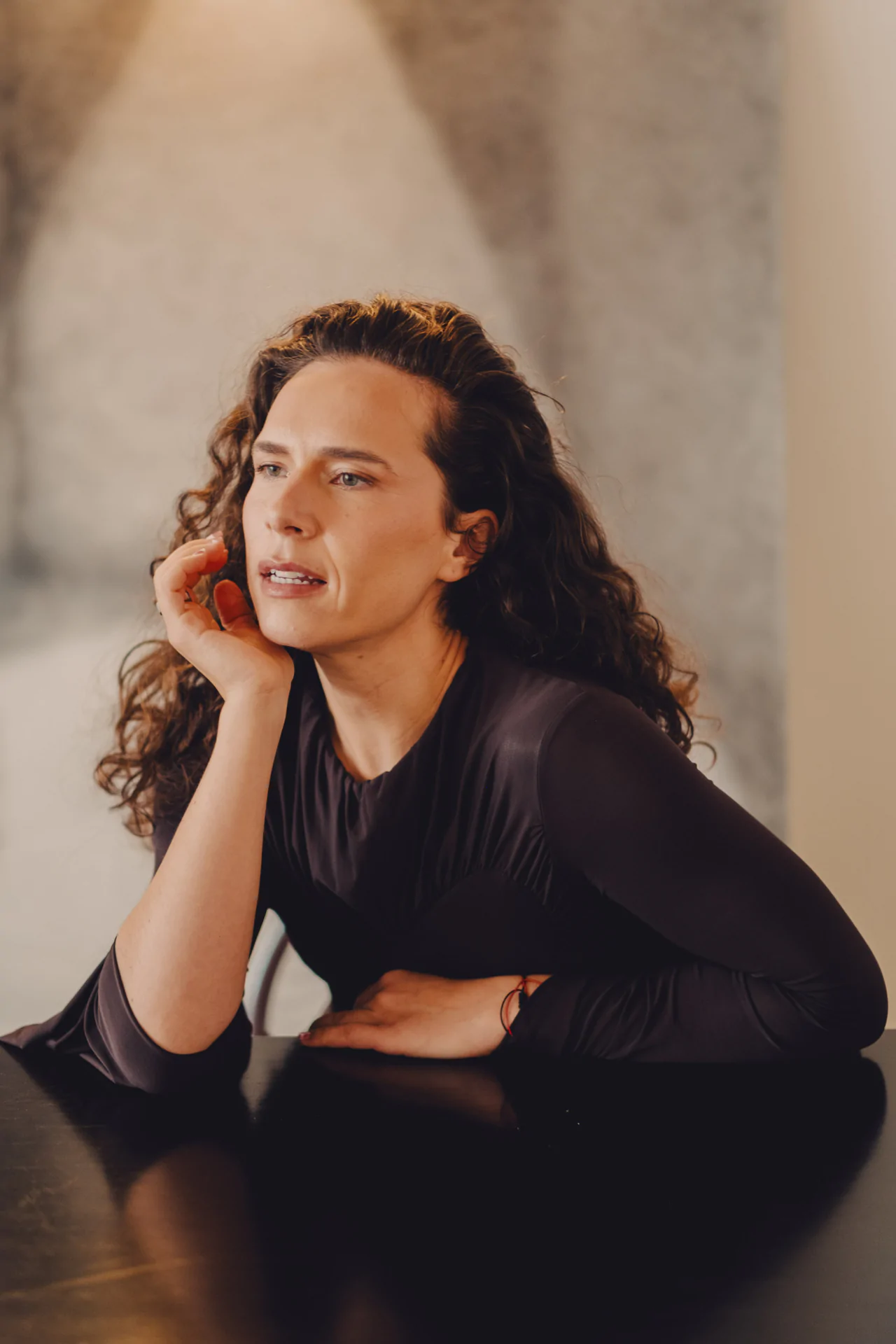 Person with long curly hair resting their chin on their hand, seated at a table, wearing a long-sleeve dark top. Neutral expression with a soft-focus background.
