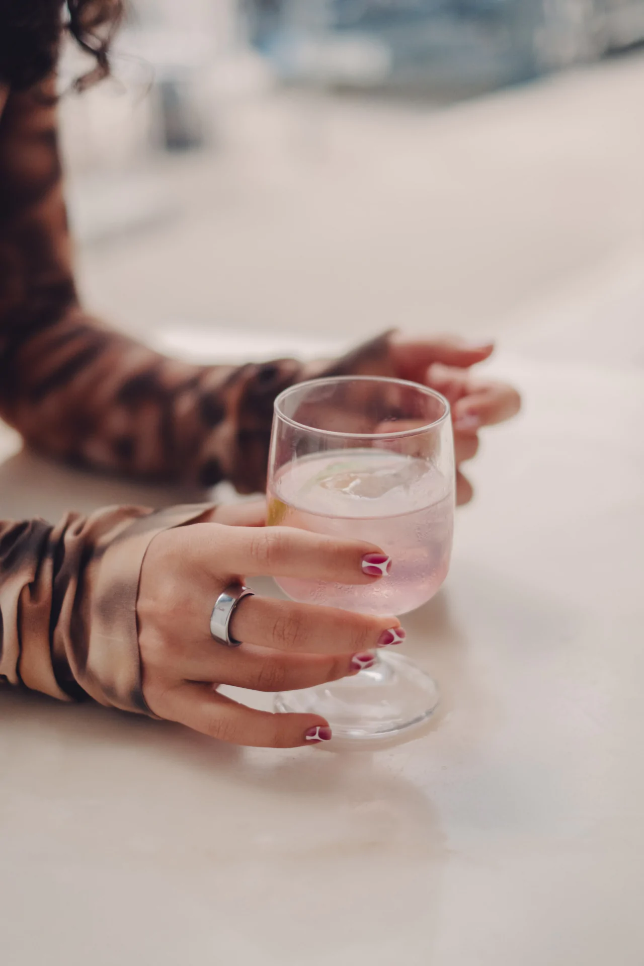 A person with painted nails holds a glass containing a pink drink with ice, sitting at a white table.