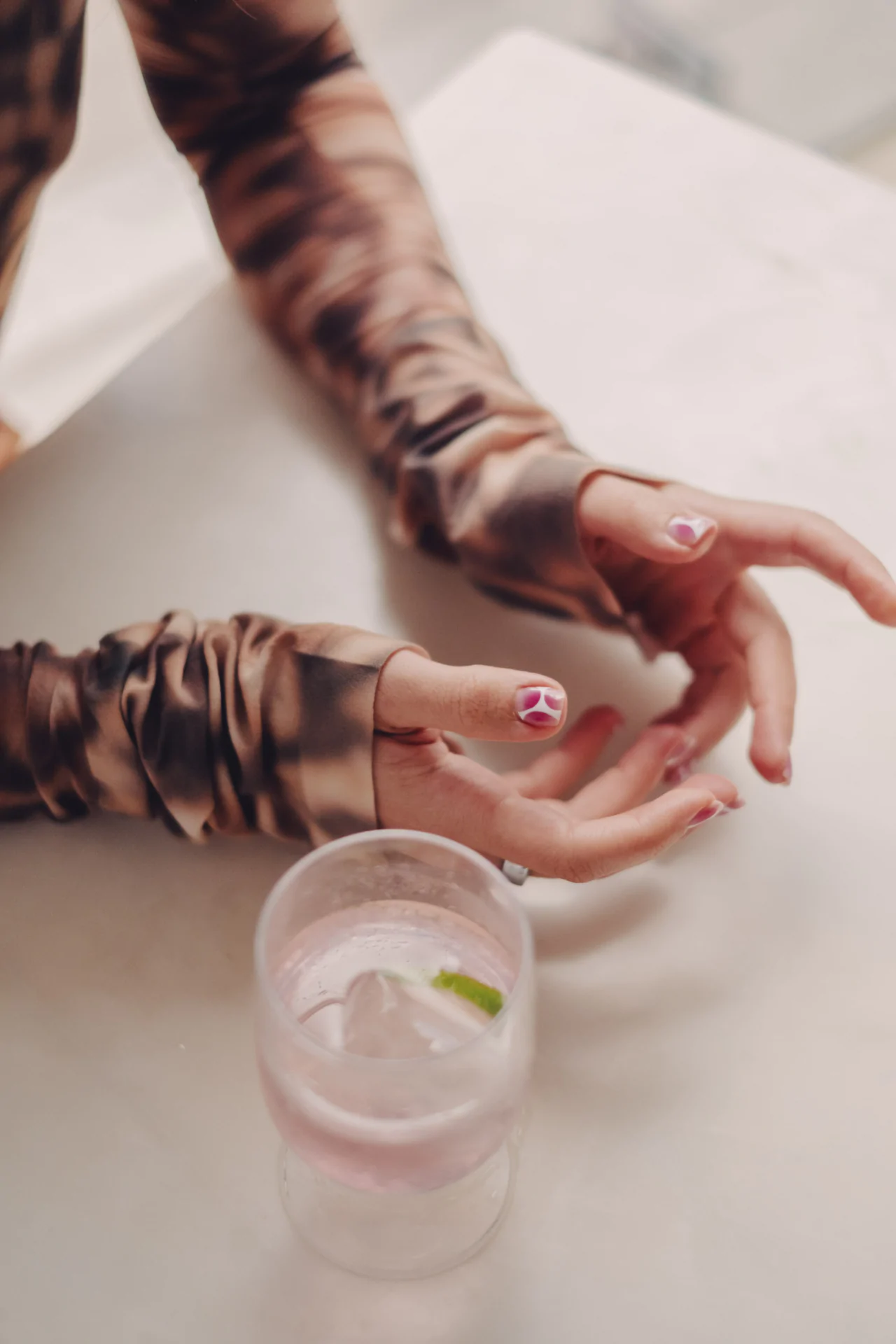 A person with long-sleeved, patterned top holds a clear drink with ice and lime on a white table. Their nails have pink and white designs.