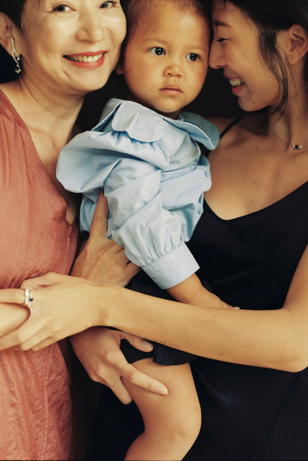 Two women smile while holding a young child dressed in a blue outfit. One woman wears a rust-colored dress, and the other a black dress. The child looks ahead with a neutral expression.
