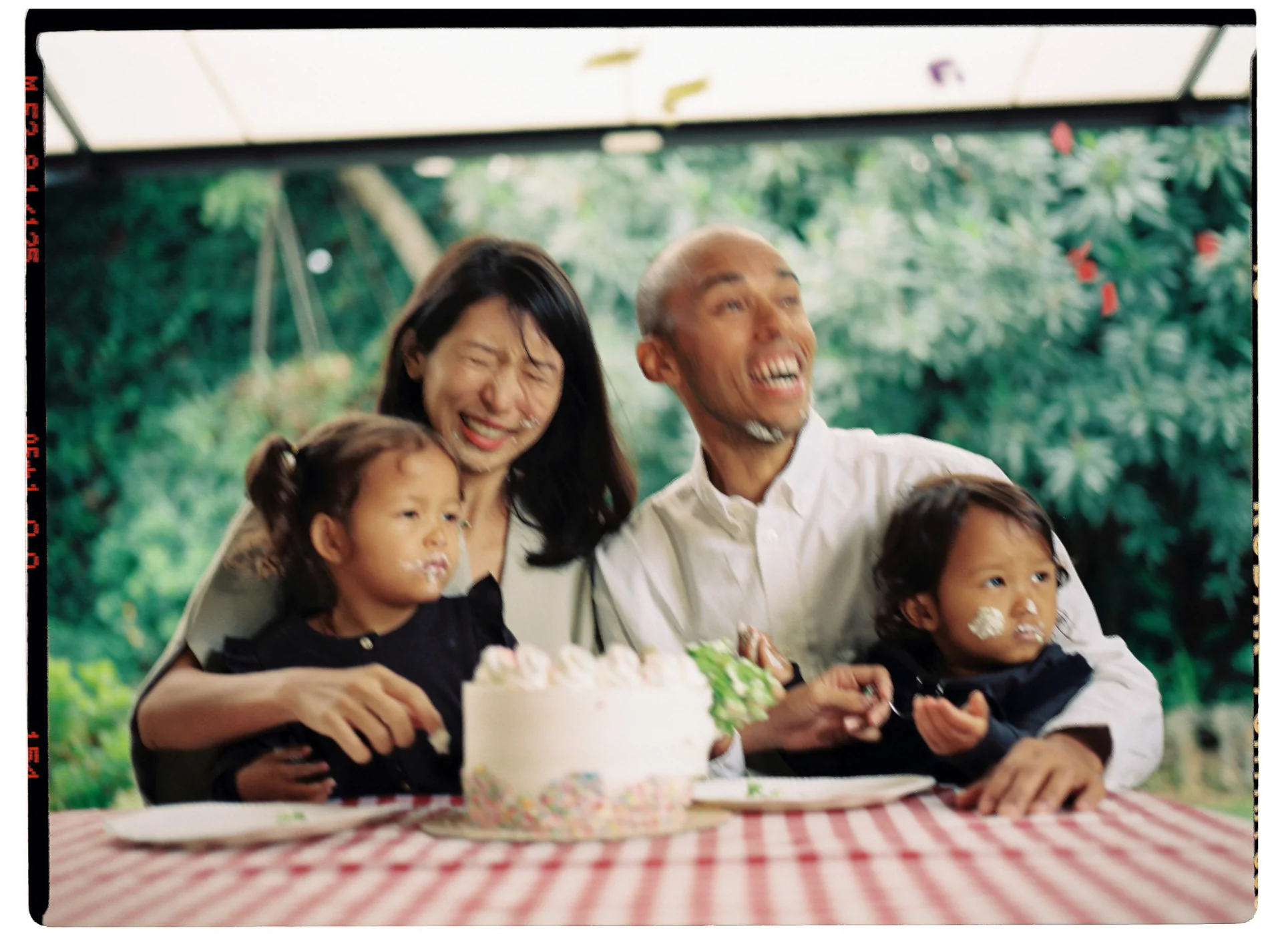 A family of four joyfully sits at a table with a cake. The two children have frosting on their faces. The setting includes a red checkered tablecloth and an outdoor backdrop of greenery.