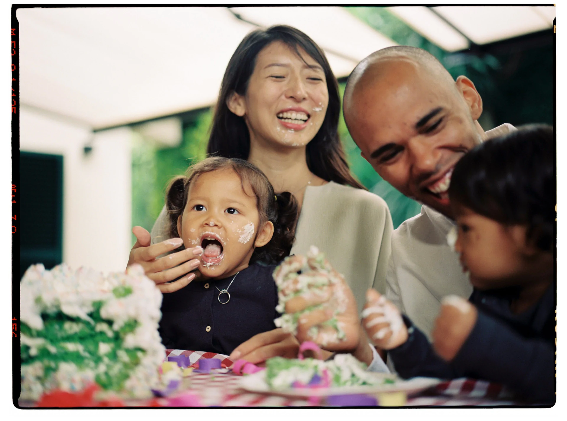 A family of four enjoys a picnic. A toddler has cake on her face and hands, while the others laugh. There is a cake on the table.