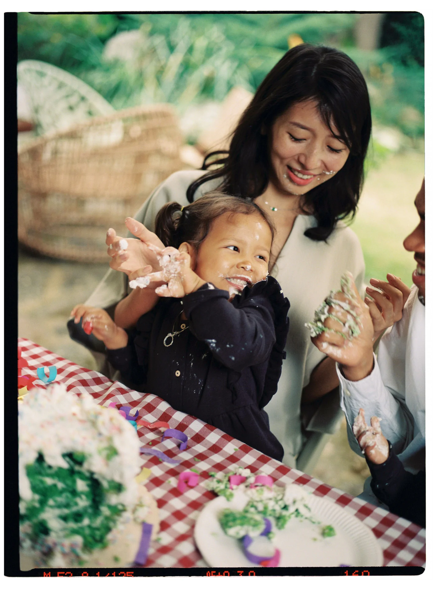 A child with cake on her face is held by an adult, seated at a red checkered table with a cake and decorations. Another person is visible, slightly out of view.