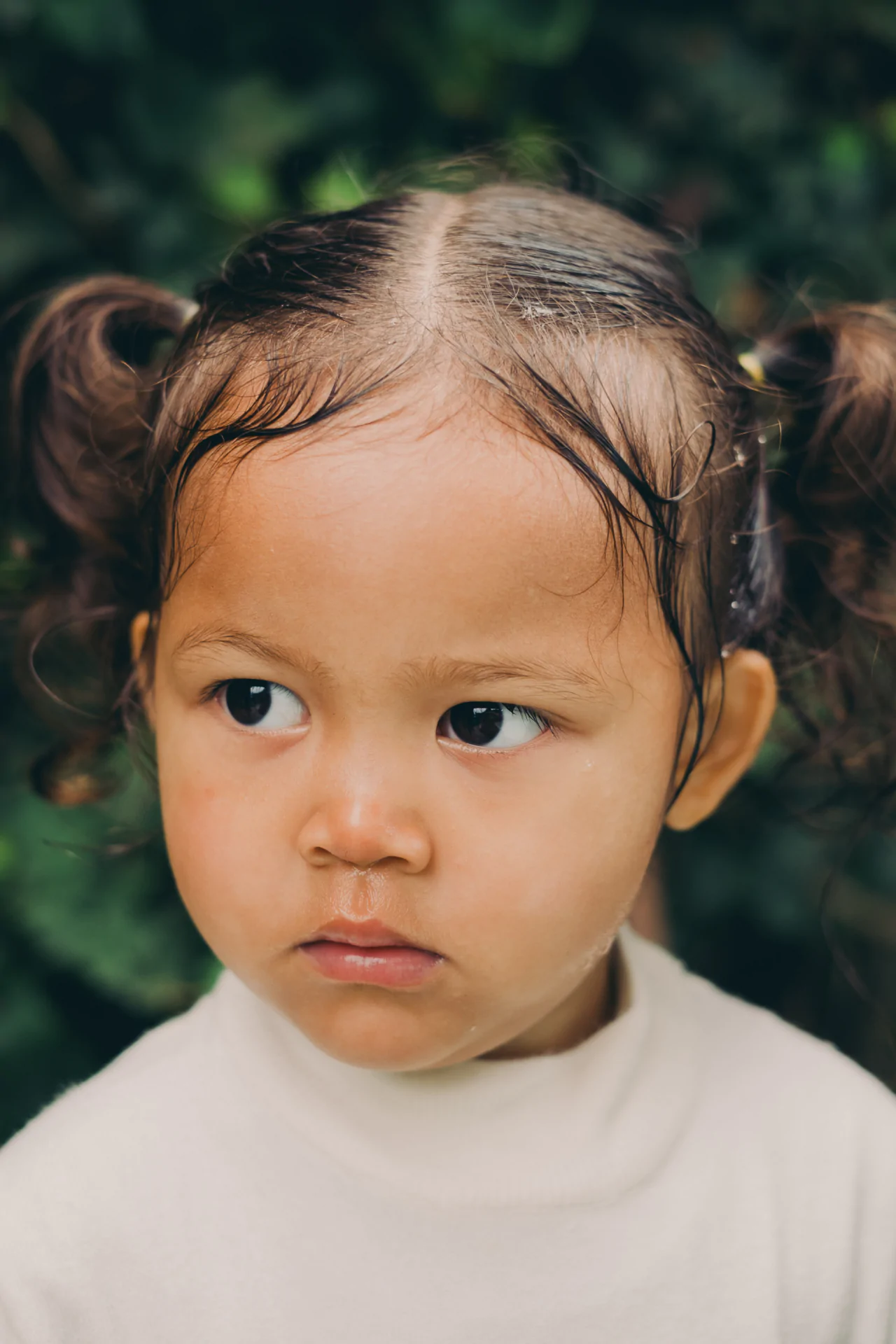 Young child with pigtails wearing a white top, looking to the side, in front of a blurred green background.