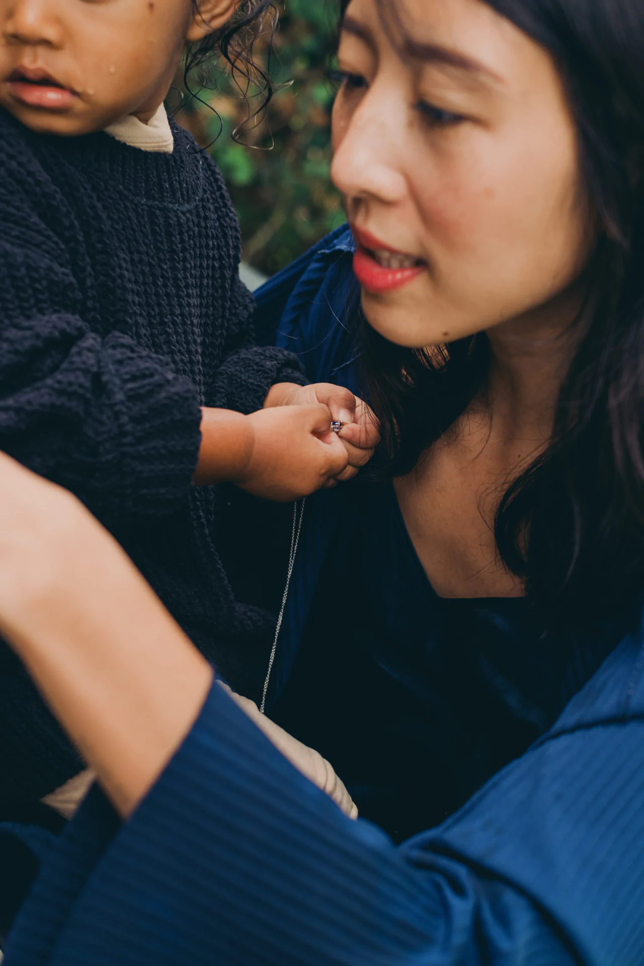 An adult and a child in dark clothing are close together outdoors. The child holds a necklace while the adult looks at them affectionately.