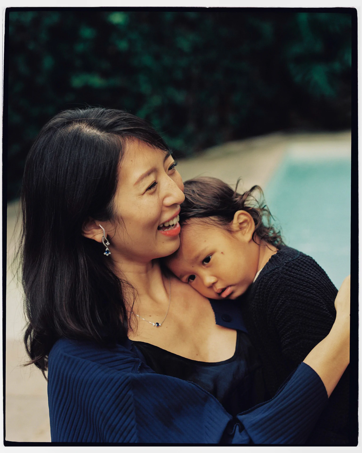 Woman holding a young child by a pool, both wearing dark clothing. The child rests their head on the woman's shoulder.