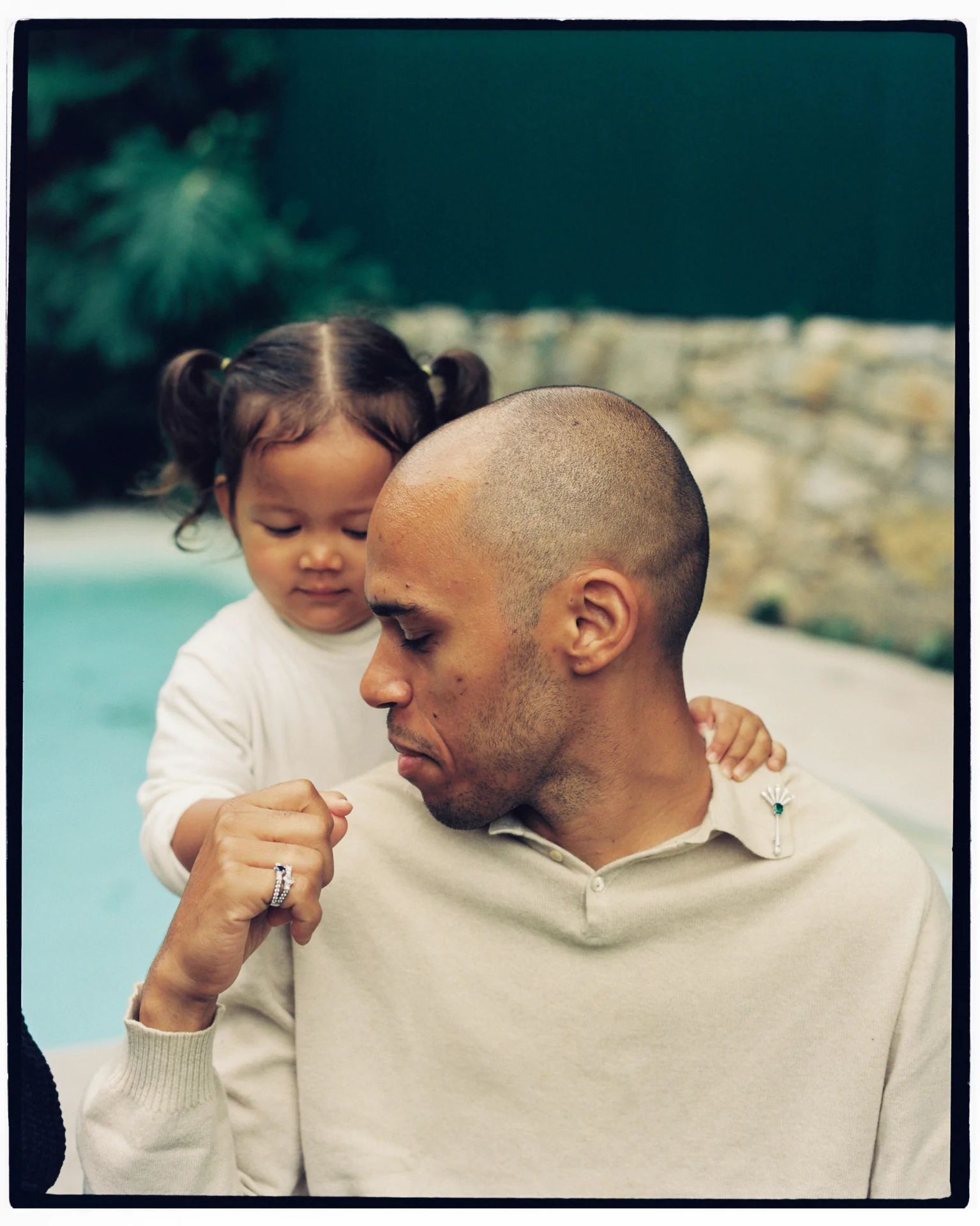 A man with a shaved head sits in front of a young child with pigtails by a pool. The child places a hand on his shoulder. They both wear light clothing.