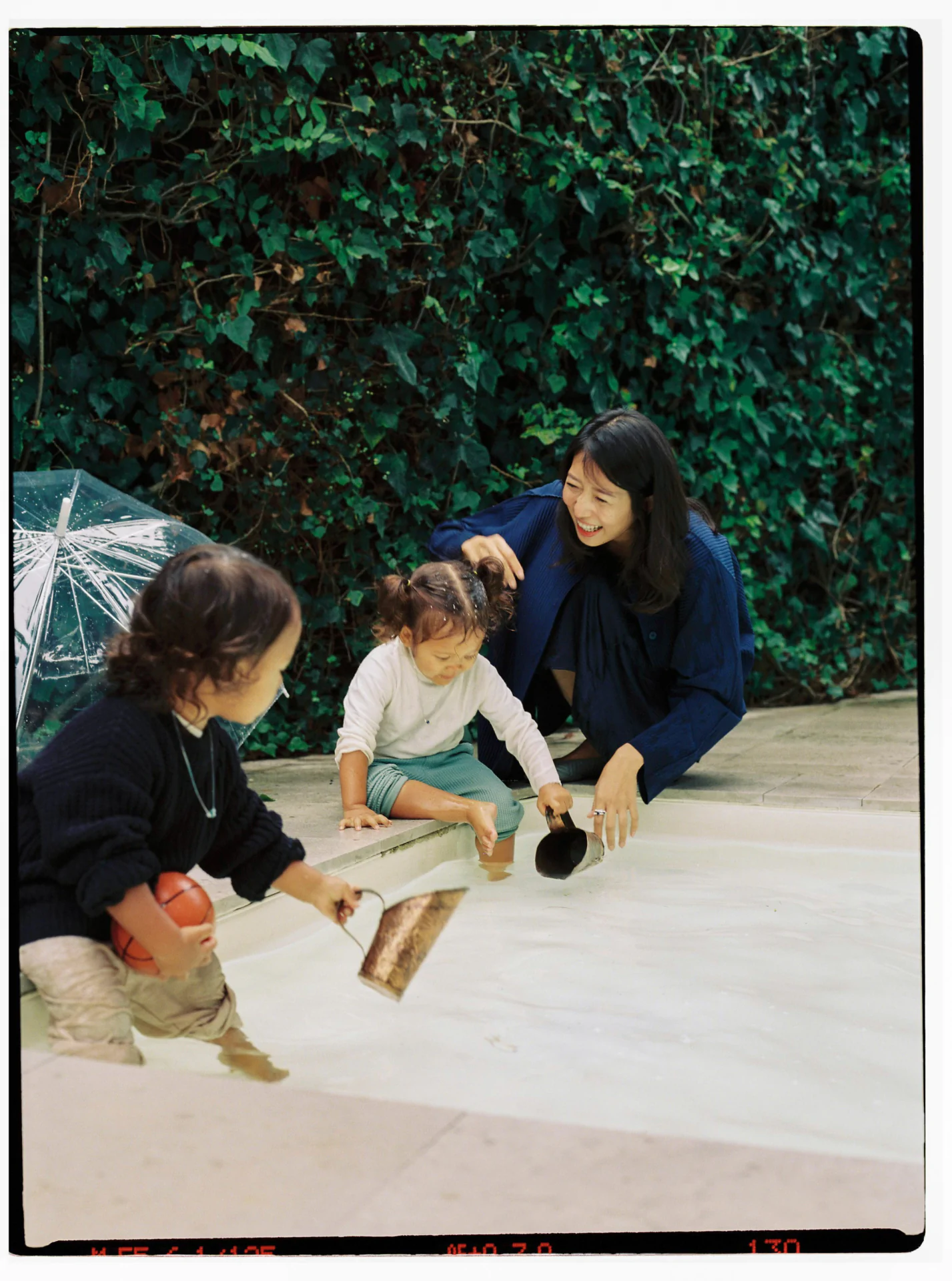 Woman and two children play by a shallow pool. One child holds a ball, the other scoops water with a tool. Lush green foliage in the background.