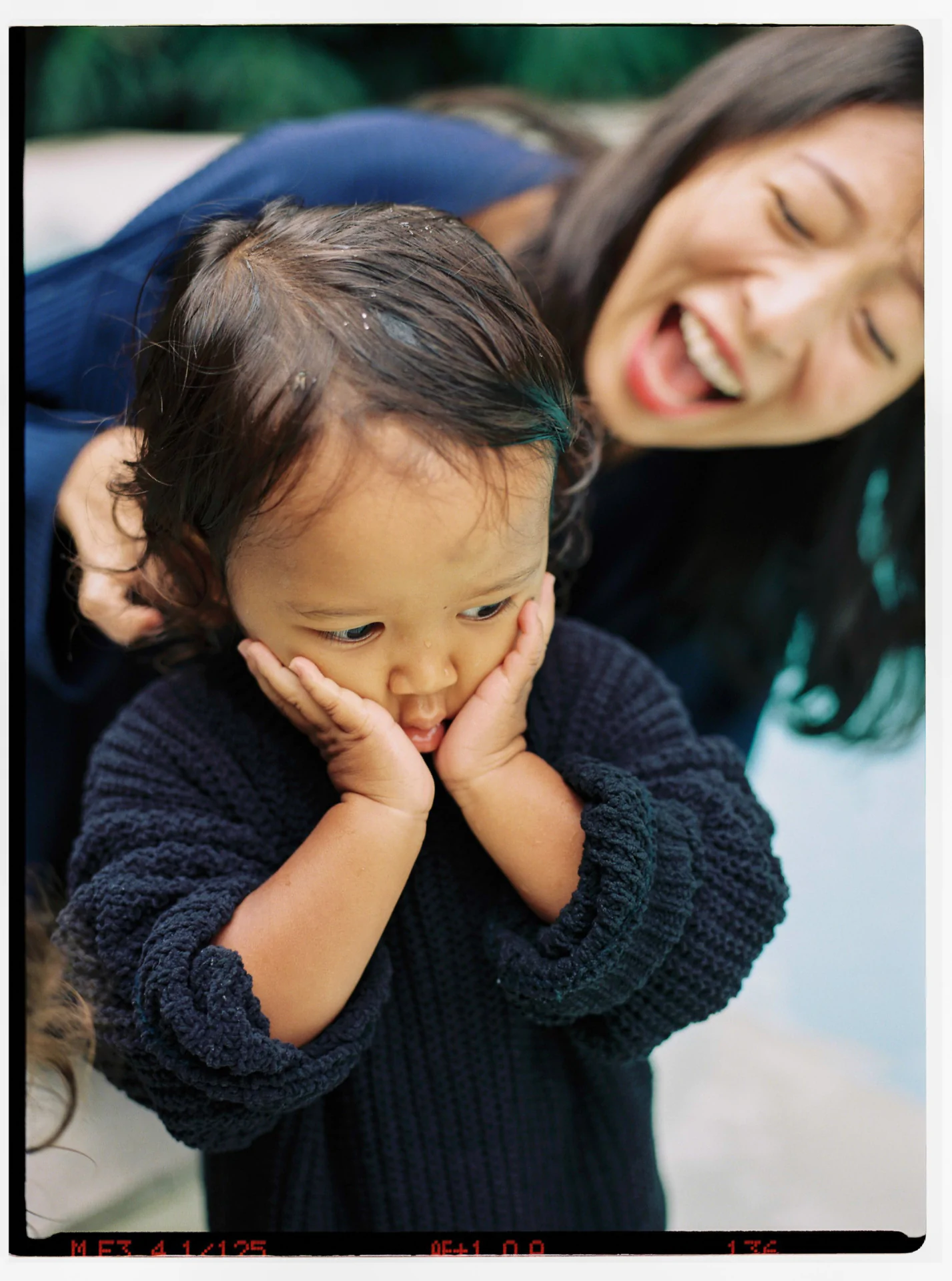 A child in a dark sweater looks pensive with hands on cheeks, while an adult in the background smiles widely.