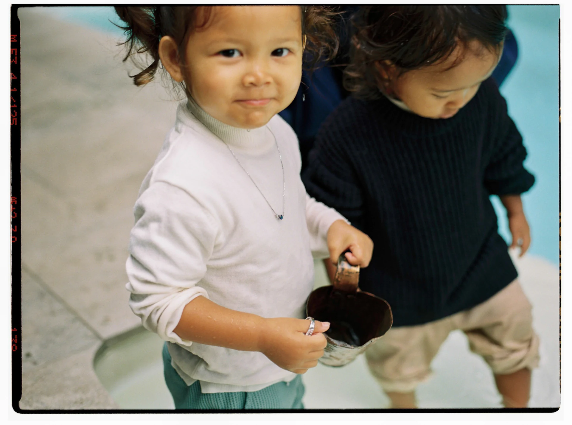 Two young children stand by a pool. One child in a white top holds a metal cup, while the other wears a black top and looks down.