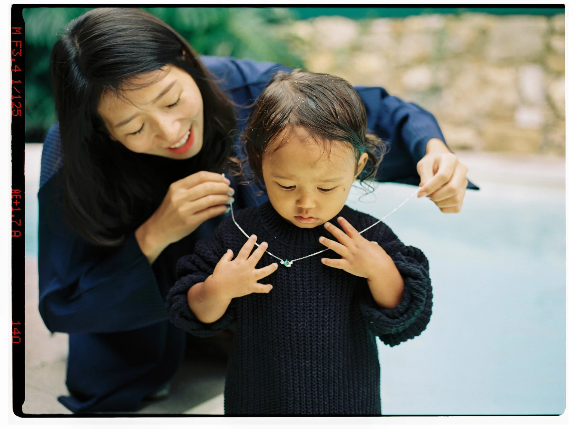 An adult helps a child wearing a knitted sweater adjust a necklace near a swimming pool.