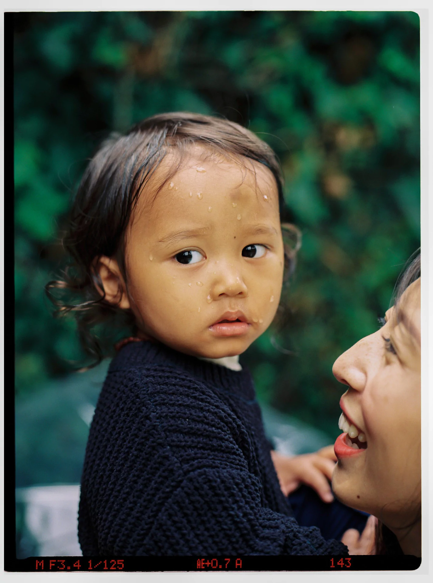 A young child with wet hair looks ahead, droplets on their forehead. An adult is beside them, smiling. Greenery is blurred in the background.
