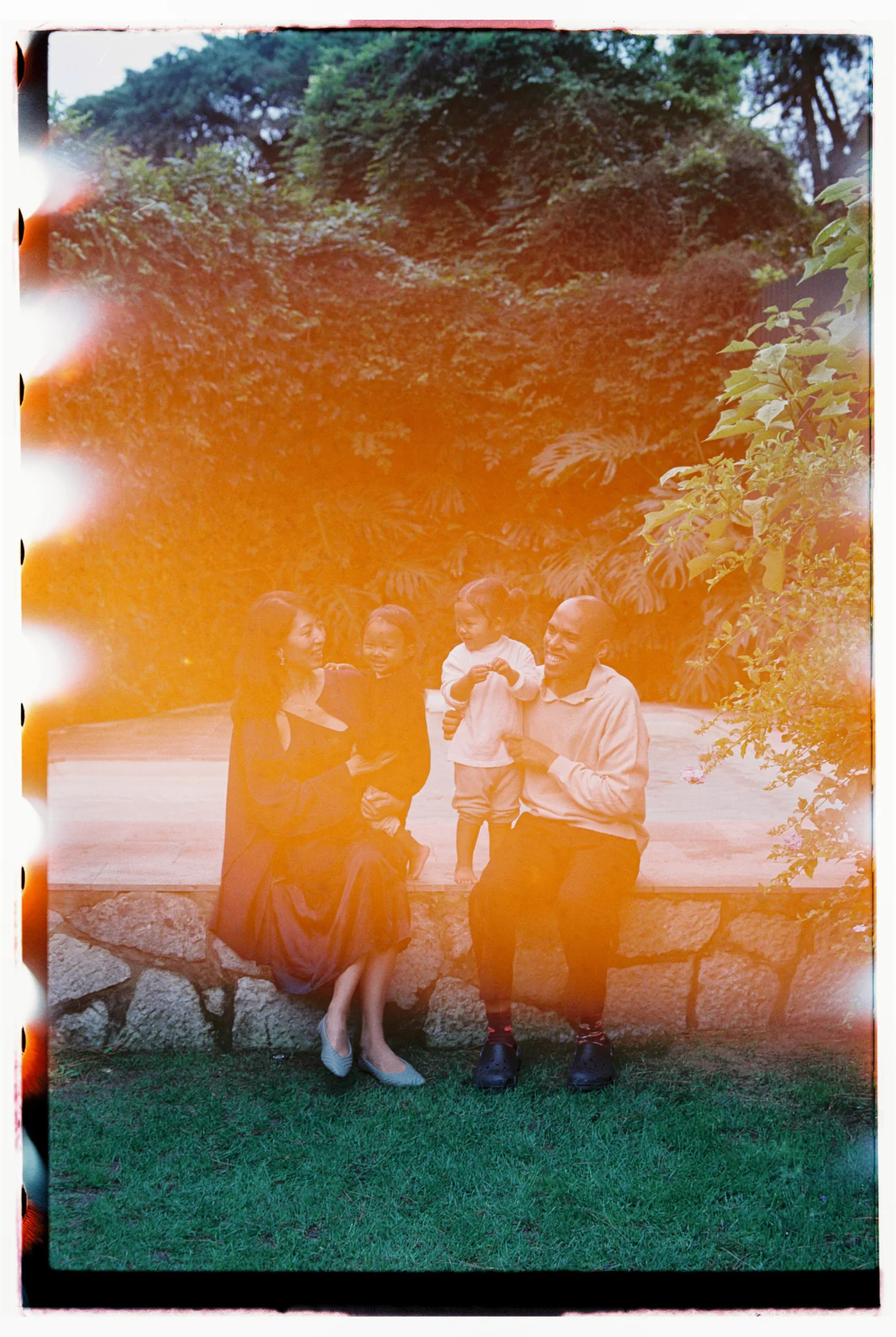 Family sitting outdoors on stone ledge in garden. Two adults and two children smiling. Photo has light leaks and a warm, vintage effect.