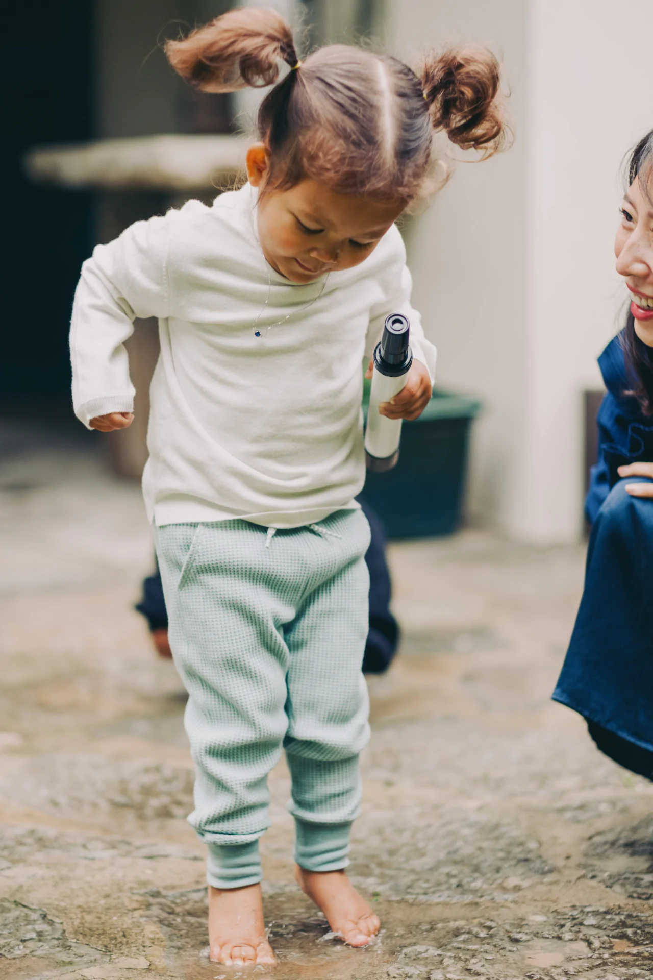 A child with pigtails wearing a white sweater and green pants stands barefoot in a puddle, holding an object. An adult kneels nearby, smiling.