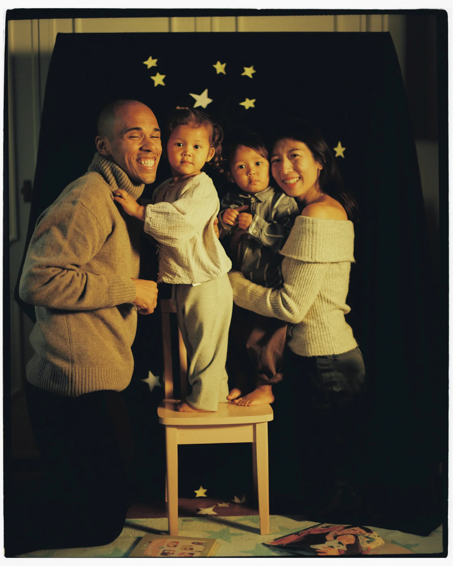 A family of four poses indoors against a backdrop with stars. Two adults hold children, one standing on a stool. A book with stars lies on the floor.