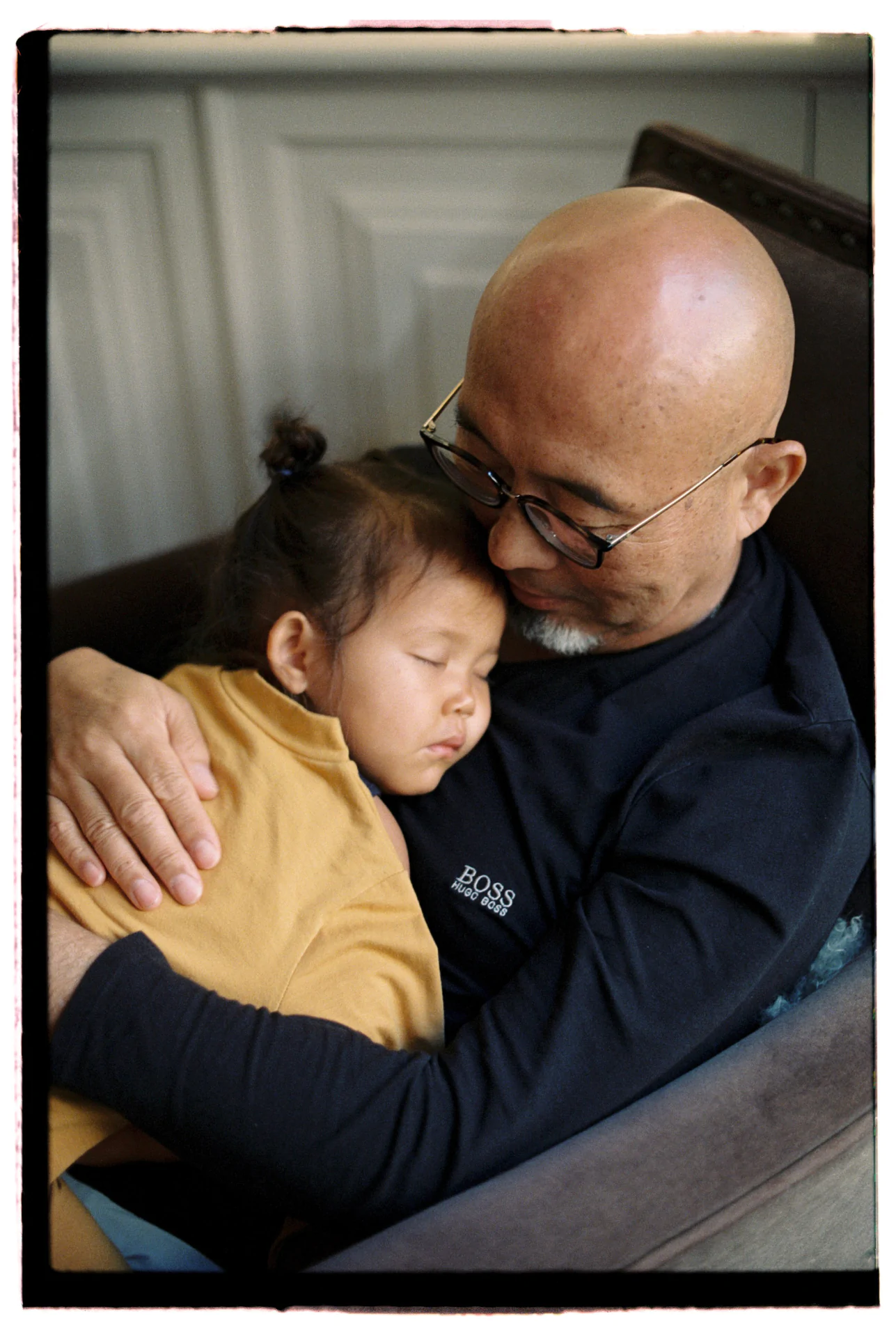 A bald man with glasses holds a sleeping child on his lap in an armchair, both appearing peaceful and resting.