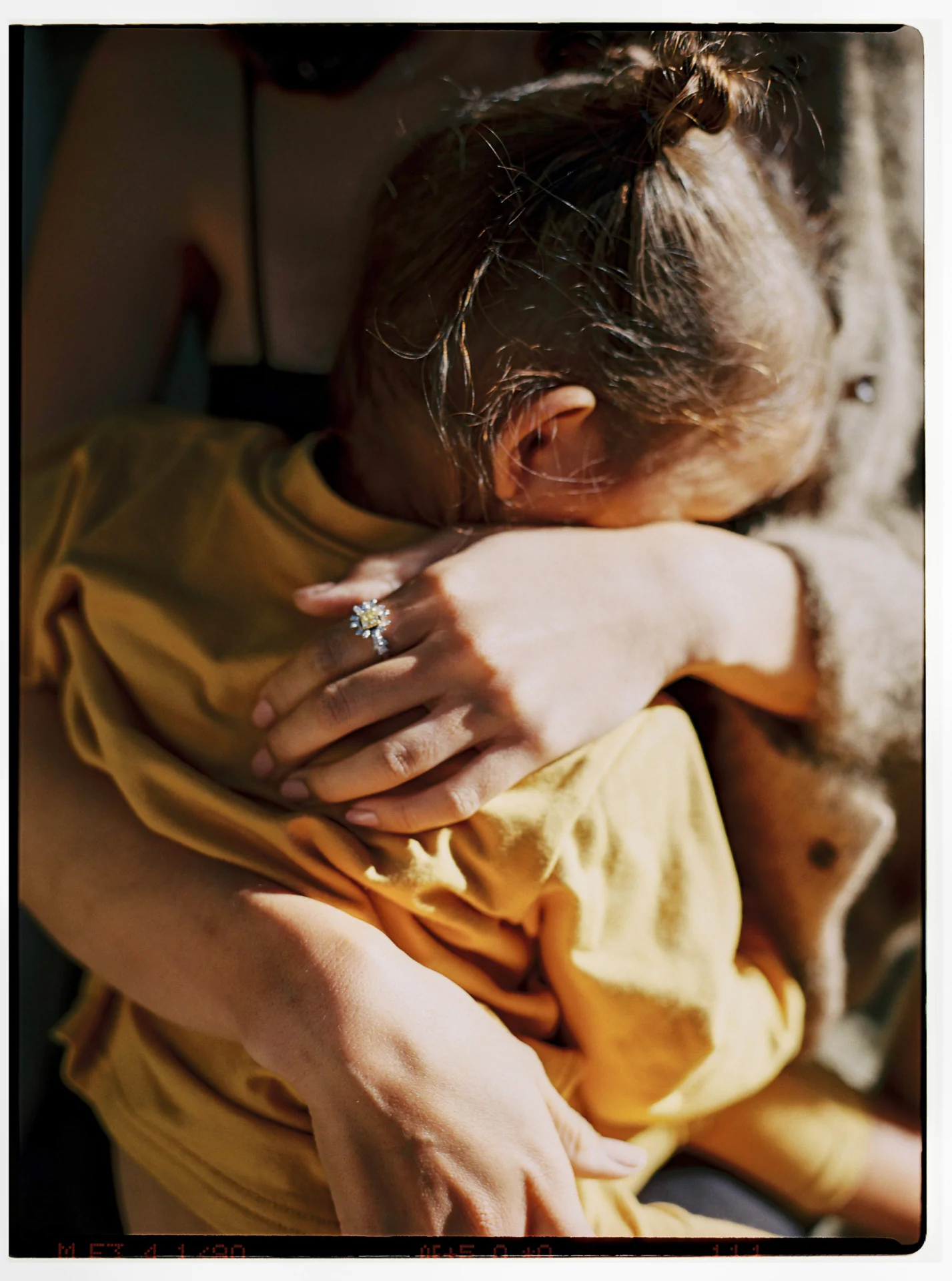 A close-up of a woman hugging a child with a topknot hairstyle. The woman wears a ring with a large stone on her finger. The child is wearing a yellow shirt.