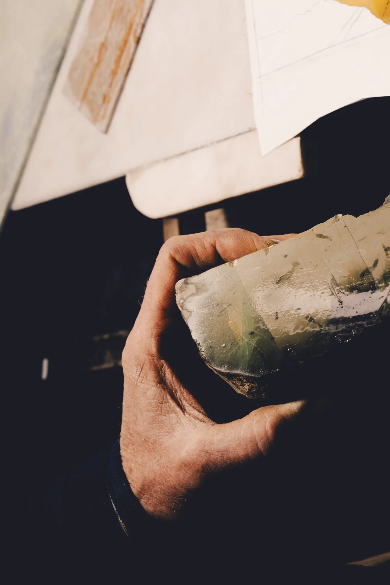 A hand holds a rough, translucent stone, with papers and a wooden surface visible in the background.