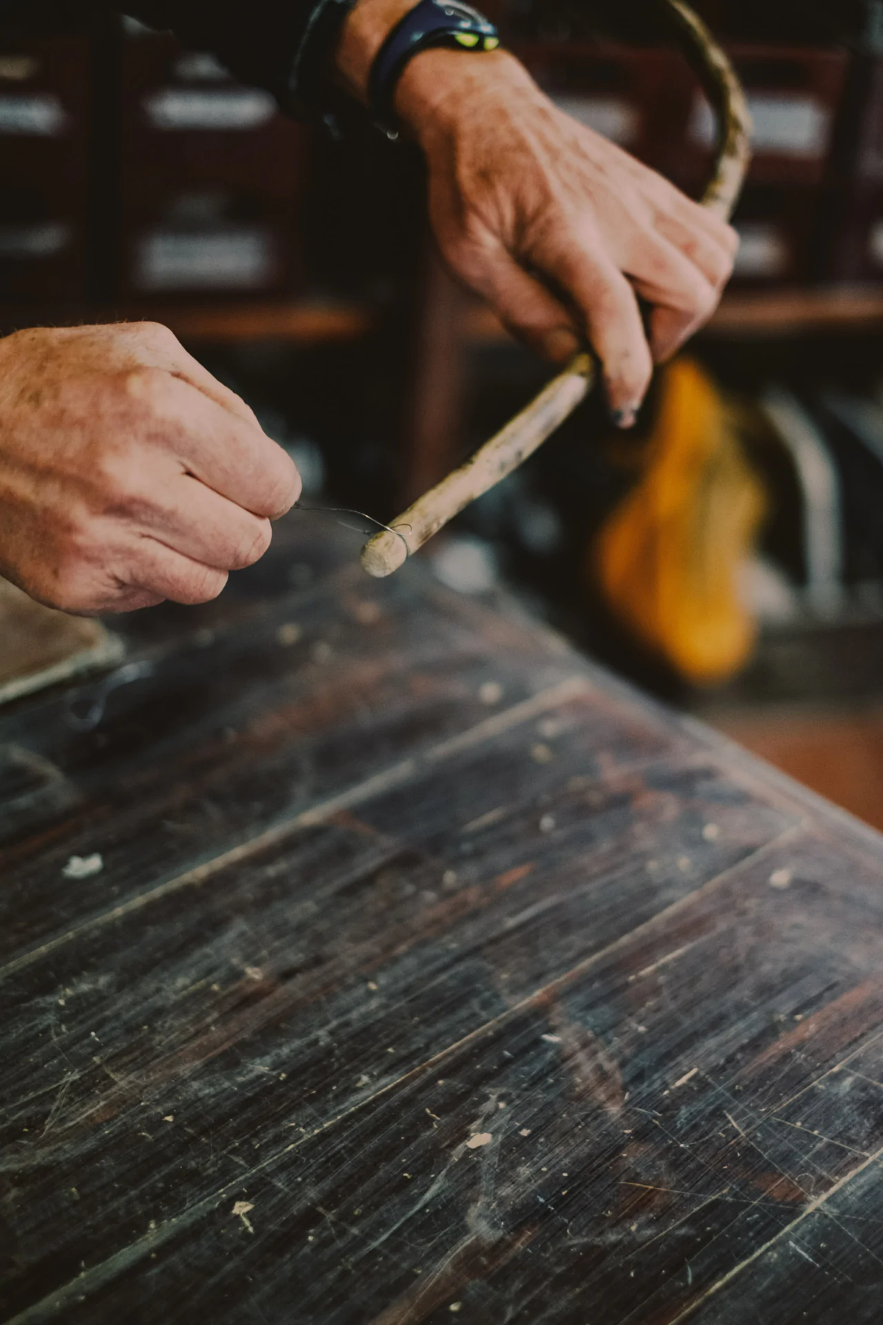 Hands bending a thin wooden branch over a dusty wooden table, with blurred background.
