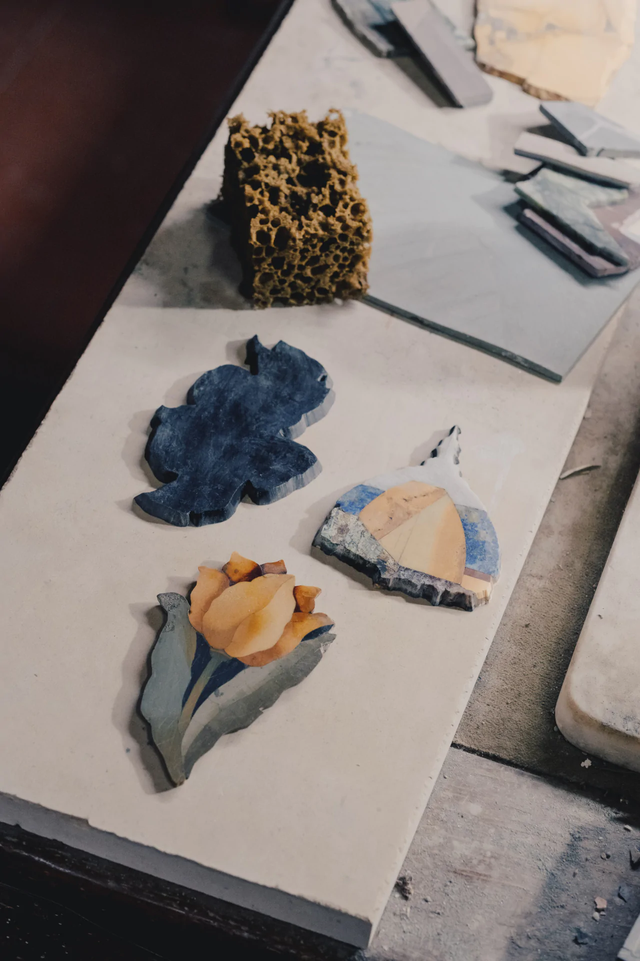 Three shaped stone pieces sit on a surface: a blue abstract shape, a dome with sky colors, and a yellow flower with blue leaves. Behind them is a porous brown sponge and other stone fragments.
