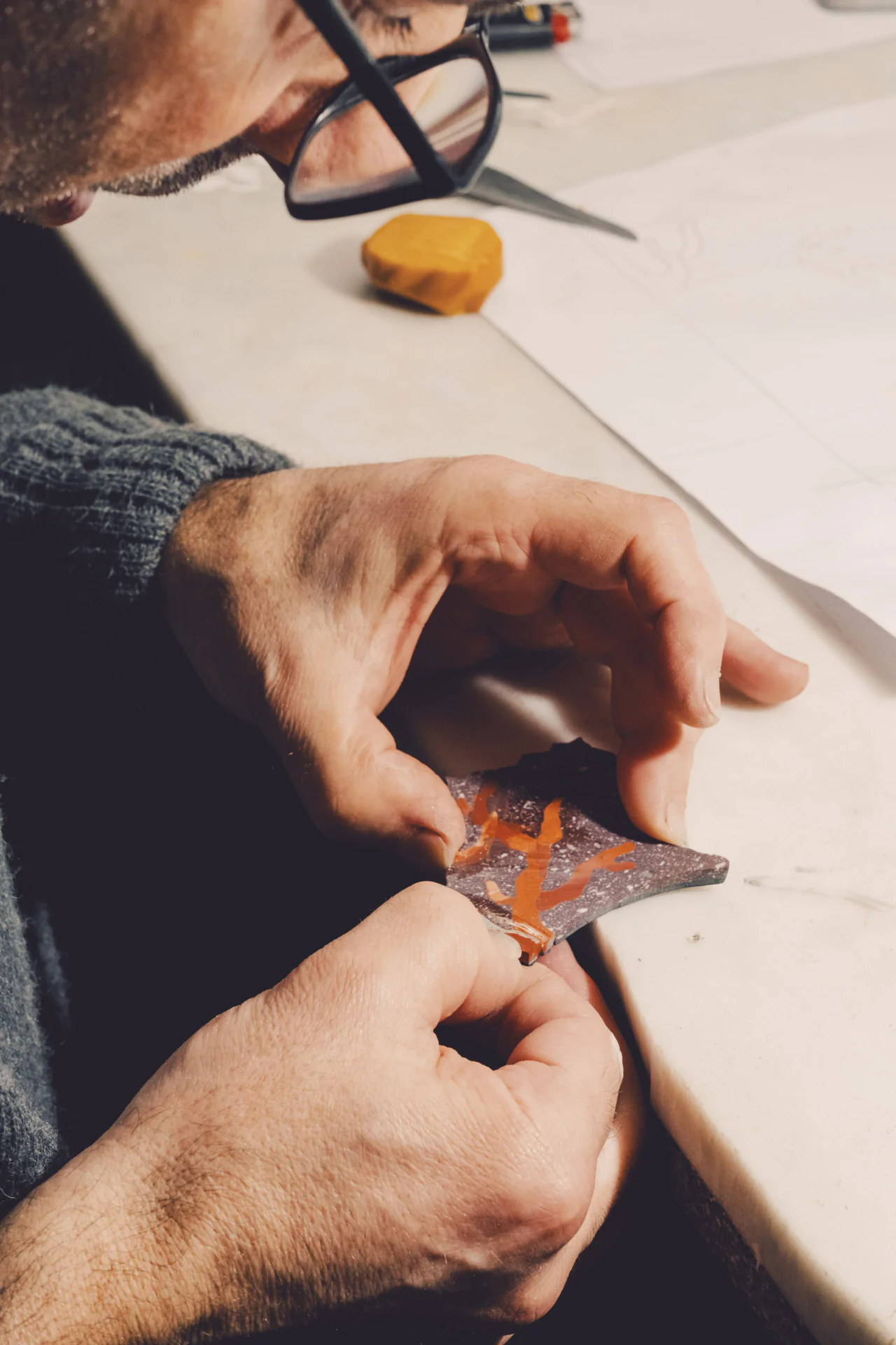 A person wearing glasses carefully paints a red design on a small, dark object at a table.