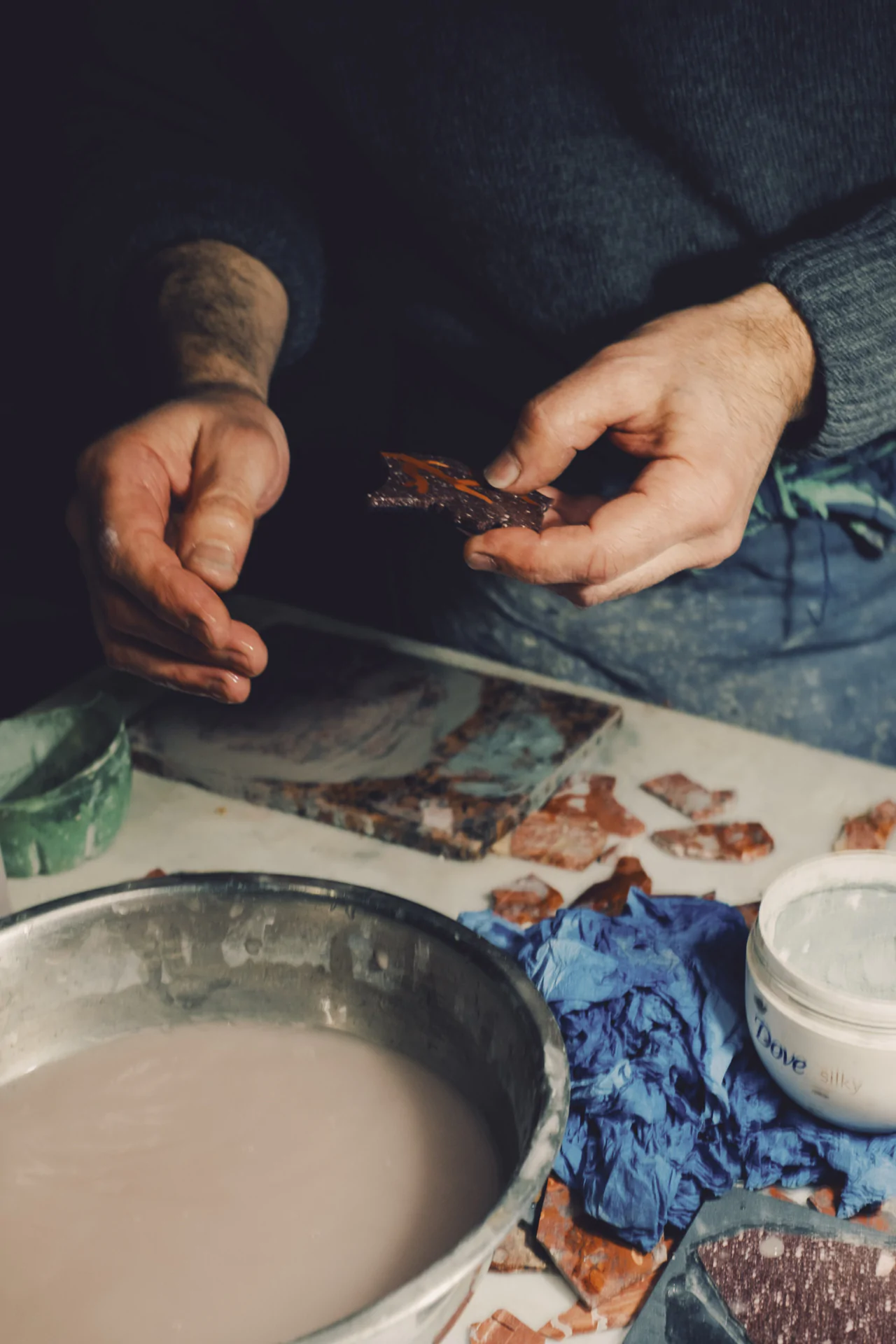 Person holding a piece of pottery over a table with clay, tiles, and a bowl of water, possibly engaged in a craft or restoration activity.