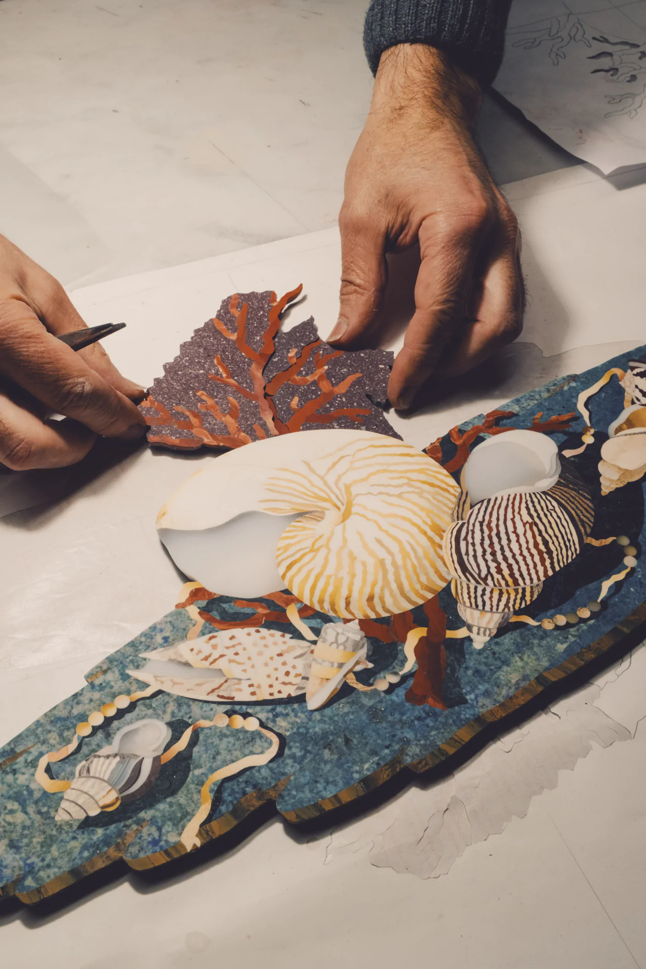 Hands assembling a decorative piece featuring seashells and coral on a blue base.