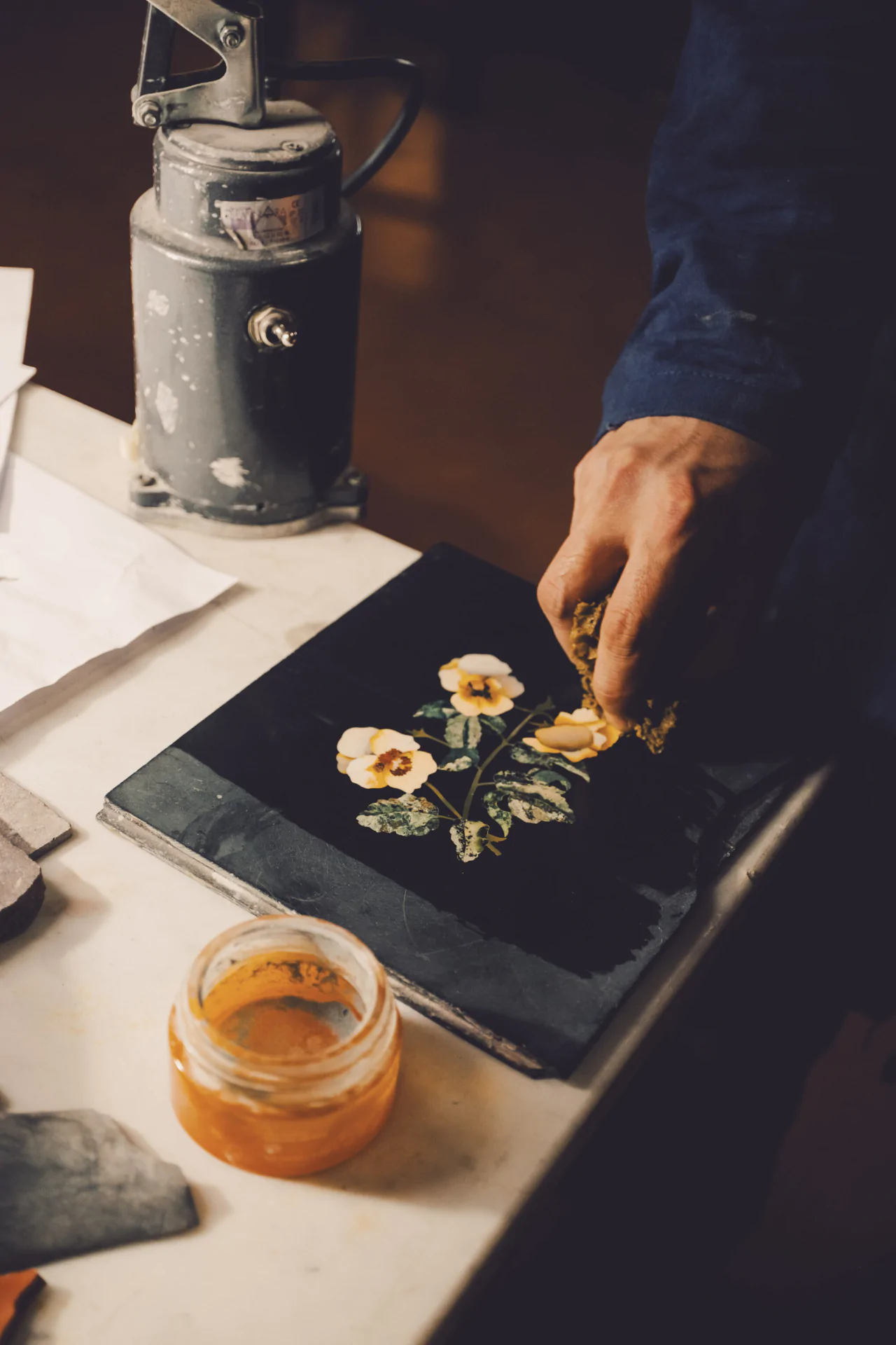 Person applying varnish to a floral design on a black surface, with a jar of yellow varnish and a mechanical press nearby.