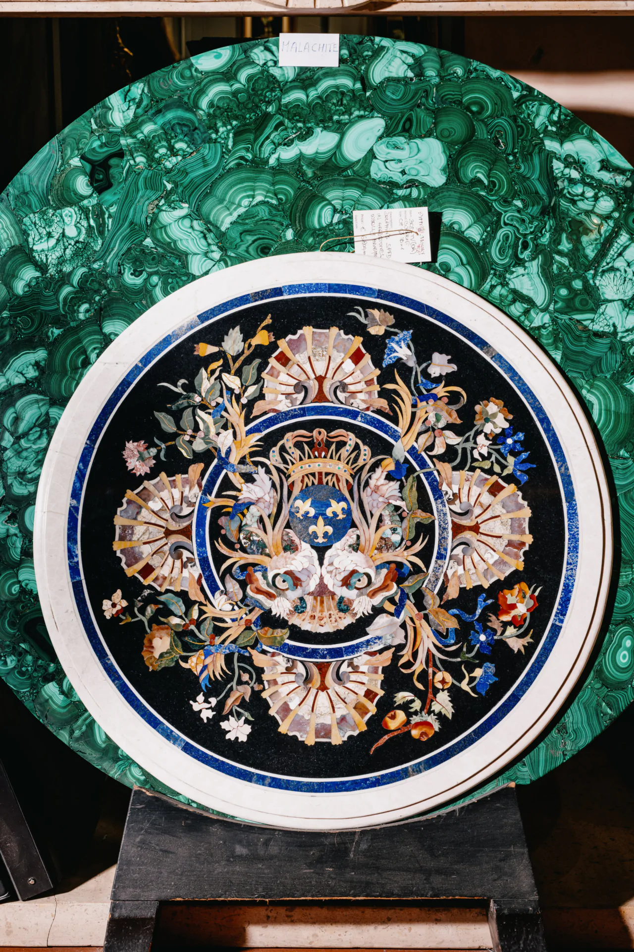 A circular artwork with an intricate floral and heraldic design on a black background, framed by a white border, with a green malachite slab in the background.