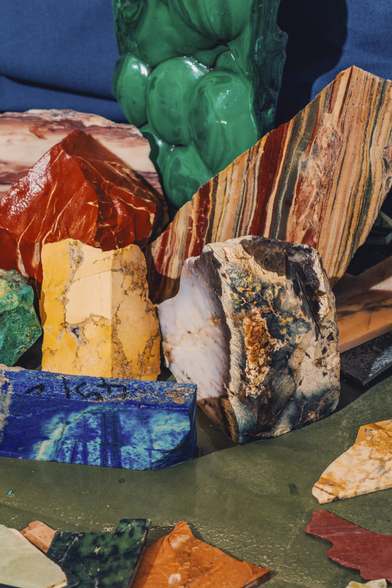A collection of variously colored minerals and stones, including green, red, yellow, blue, and striped patterns, arranged on a green surface.