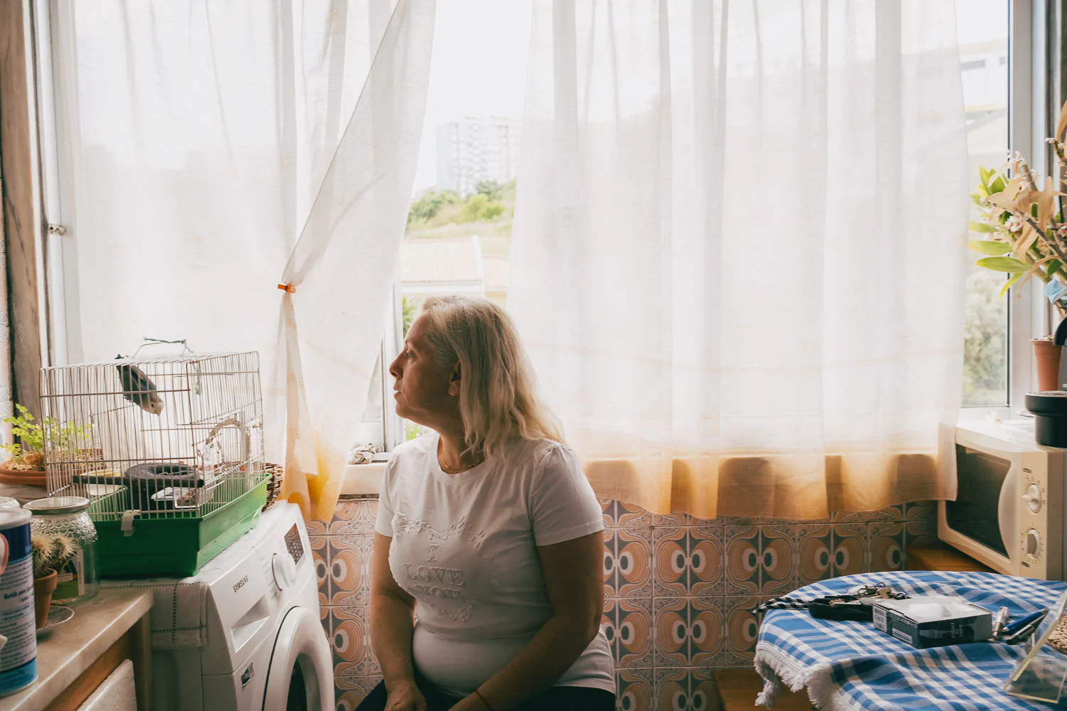 An older woman sits by a window in a sunlit kitchen, looking outside. A birdcage, plants, and household items are visible around her.
