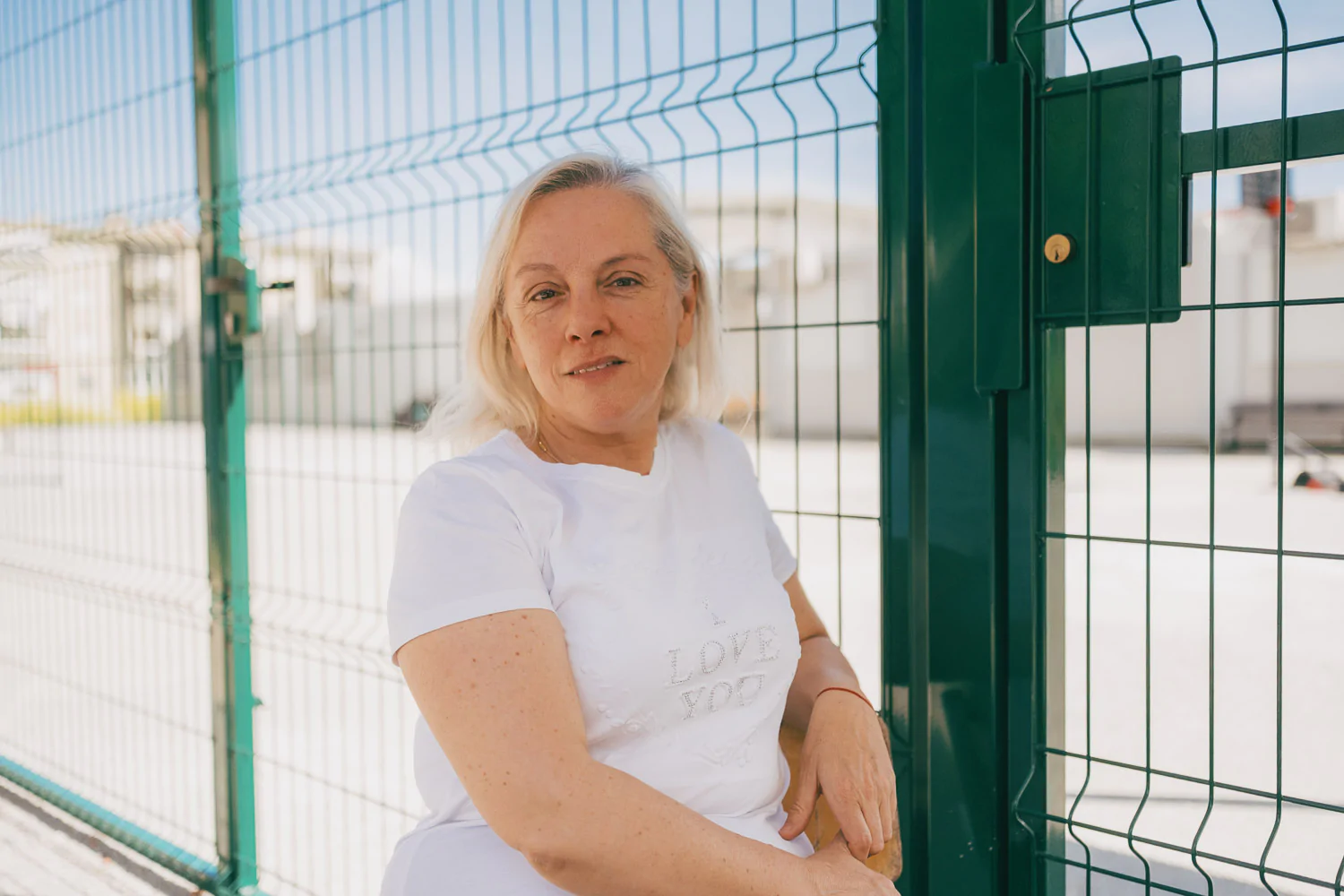 A woman with light hair wearing a white t-shirt sits by a green metal fence outdoors on a sunny day.