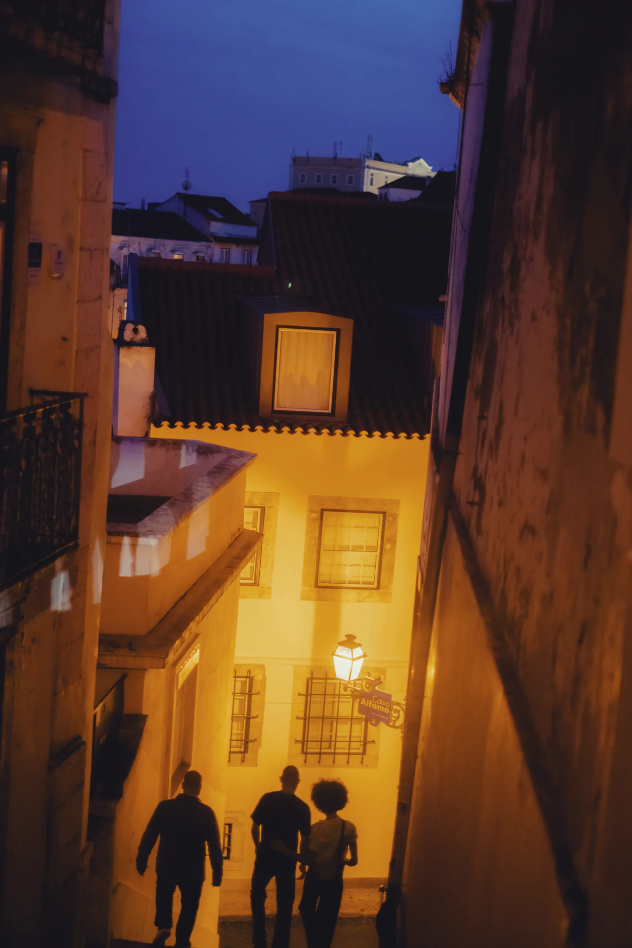 Three people walking toward illuminated yellow building at dusk Lisbon Portugal