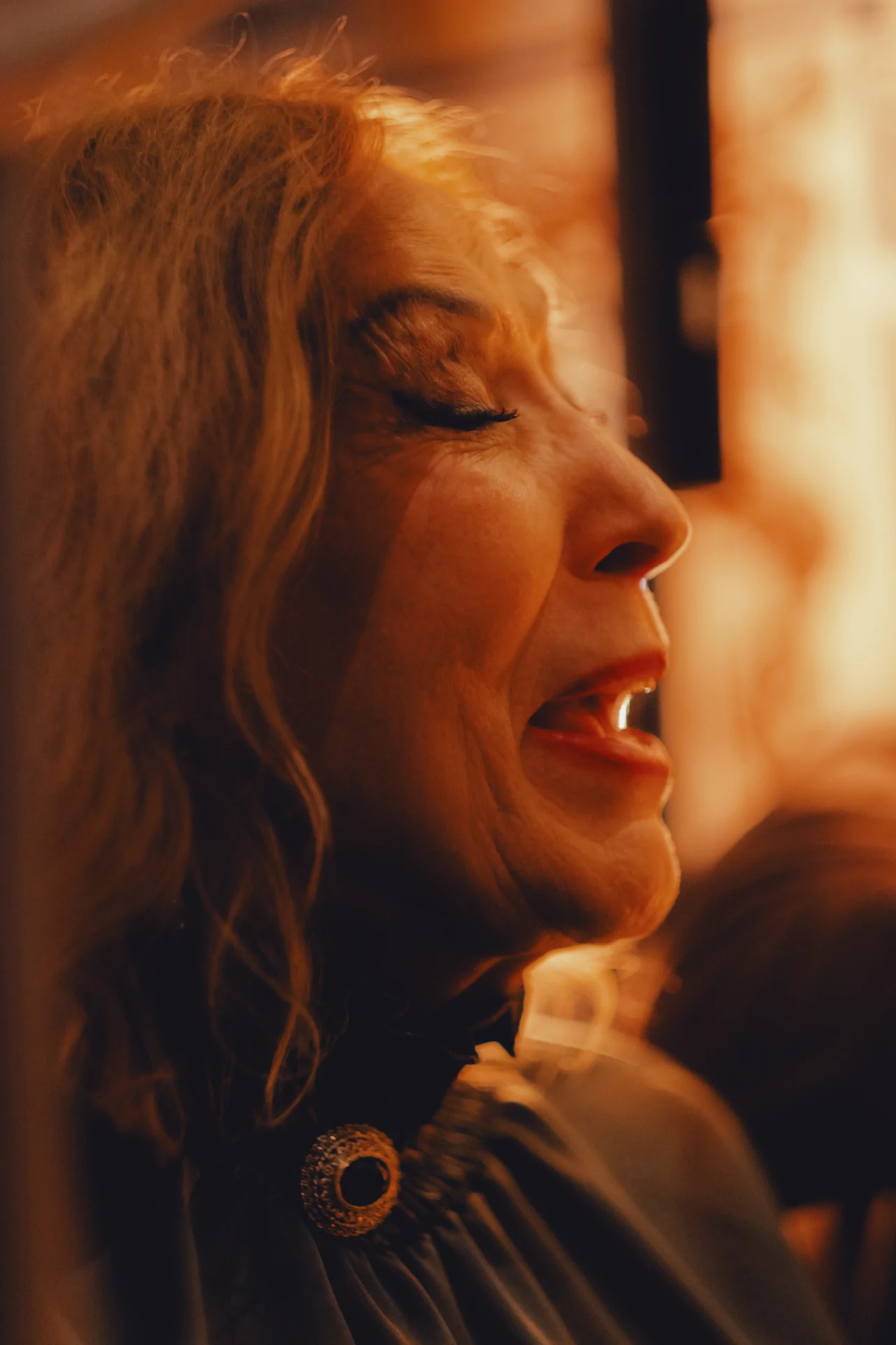 An older woman with long hair and closed eyes appears to be singing or speaking, illuminated by warm, golden light.