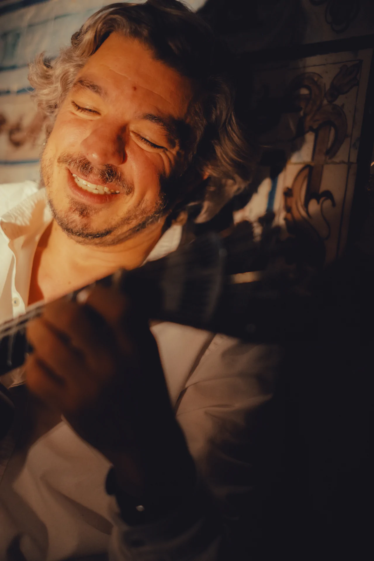 A man with gray hair and a beard smiles with his eyes closed while playing a guitar, wearing a white shirt in a warmly lit setting.