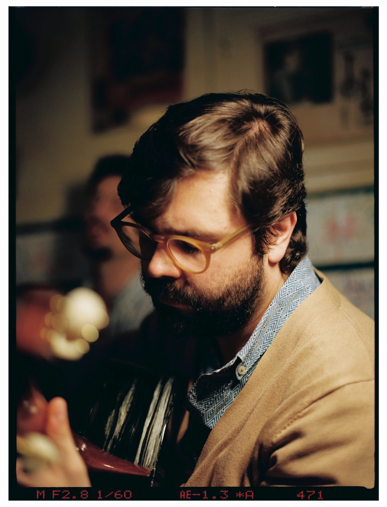 A bearded man with glasses plays a portuguese guitar indoors, with another person blurred in the background.