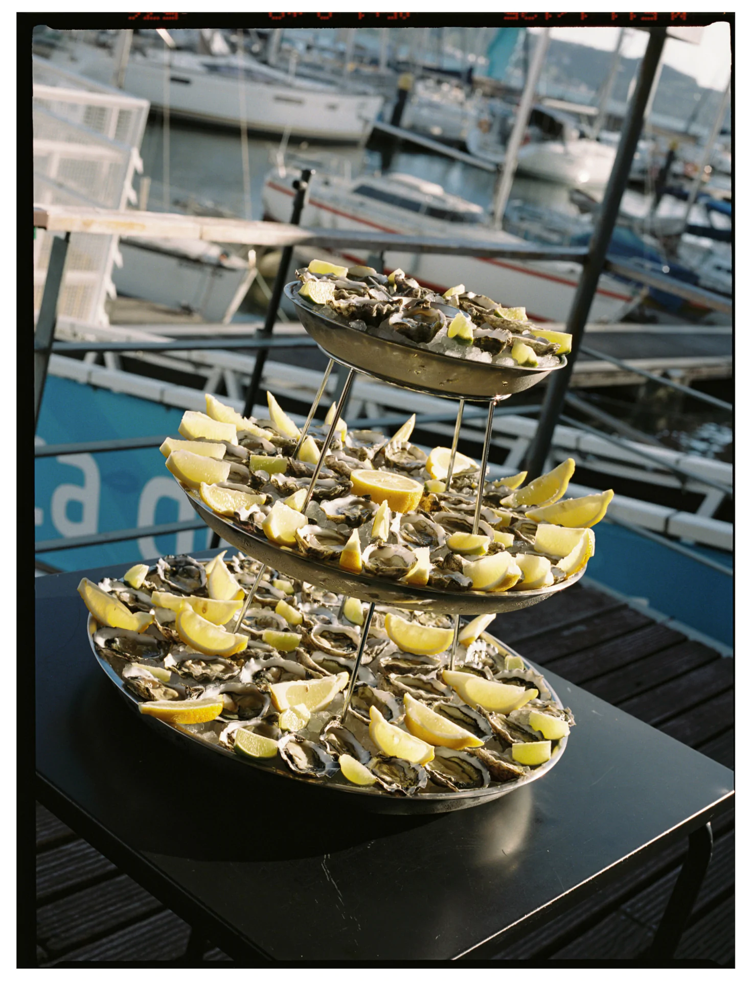 A three-tiered platter filled with opened oysters and lemon wedges sits on a table outdoors near a marina with boats in the background, in lisbon.