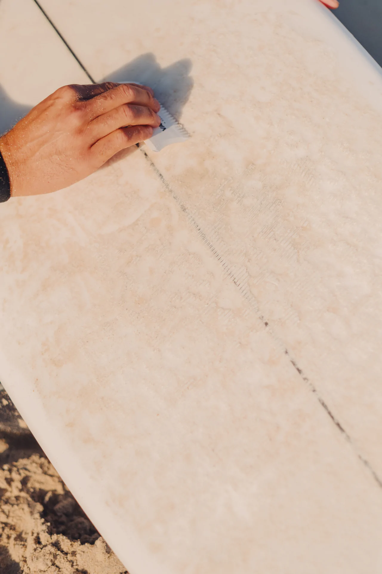 A person’s hand applies wax to the surface of a white surfboard on the sand.